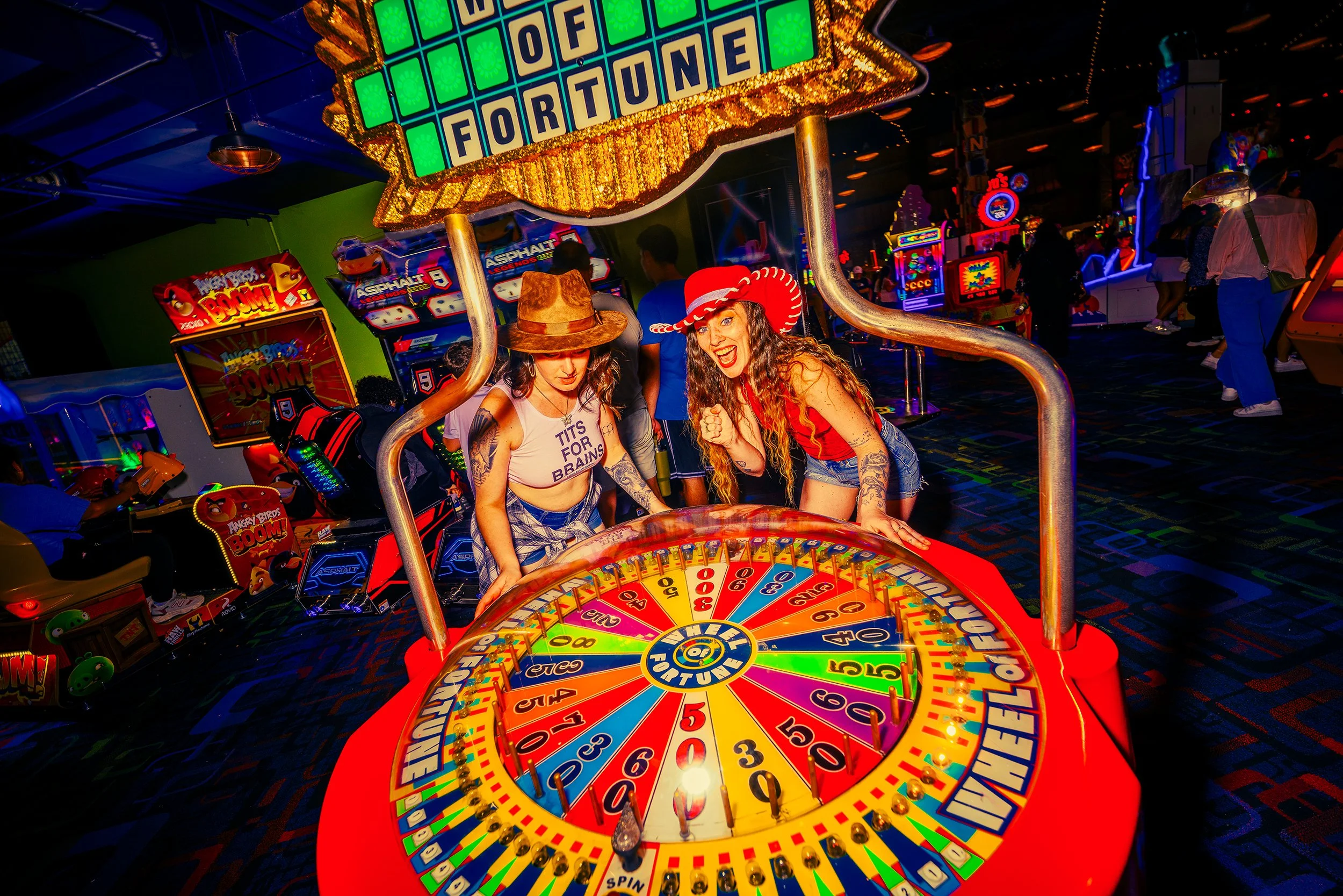 Women at a carnival game booth, one wearing a cowboy hat and the other a red and white nurse hat, playing a spinning wheel game with colorful sections and the word "Fortune" in the center.