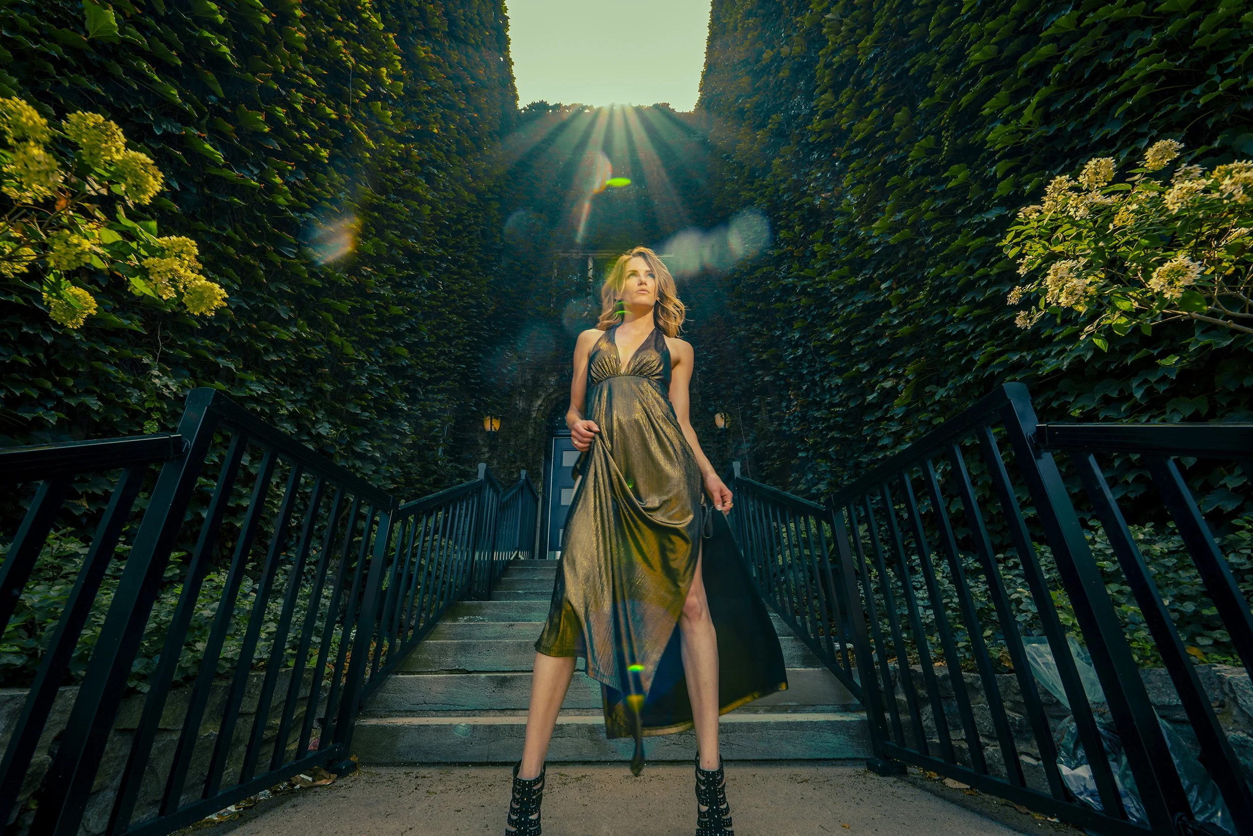 A woman wearing a metallic gold dress and high heels stands on a staircase surrounded by tall, lush green hedges. Sunlight streams down from the sky, creating lens flare effects.