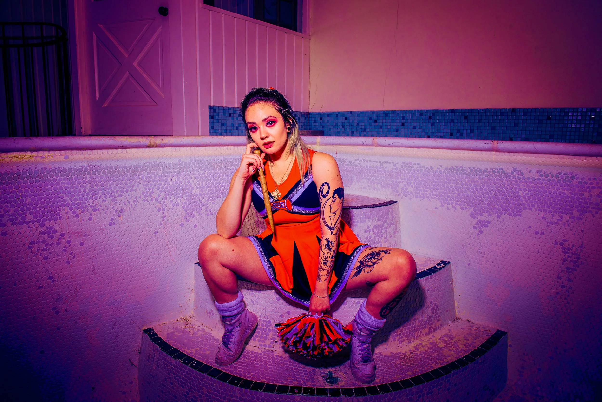 A young woman dressed as a cheerleader sitting on the edge of a small, empty pool with pink and purple lighting. She is wearing a cheerleading uniform, holding pom-poms, and has tattoos on her arms. The background features a door and tiled walls.