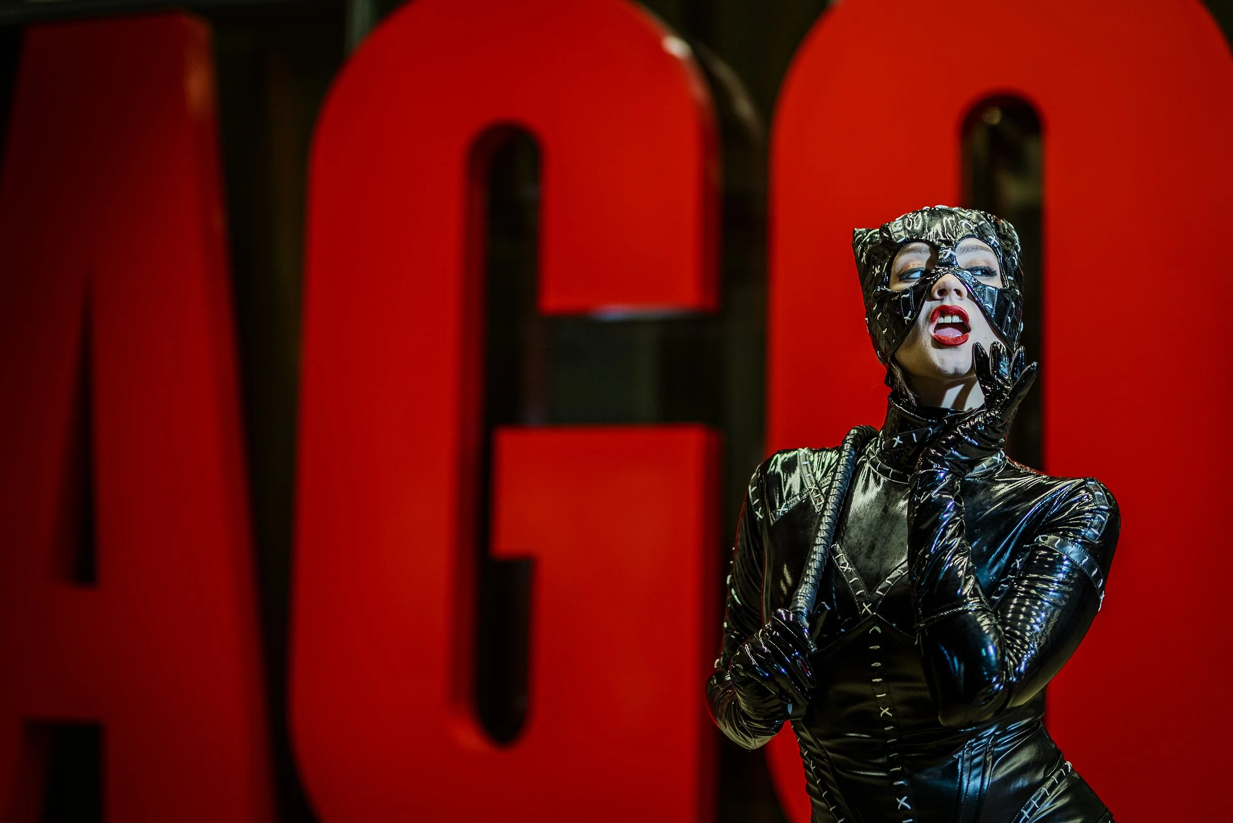 A person wearing a shiny, metallic black outfit and a matching mask, posing with their hand near their face against a backdrop with large red and black lettering.