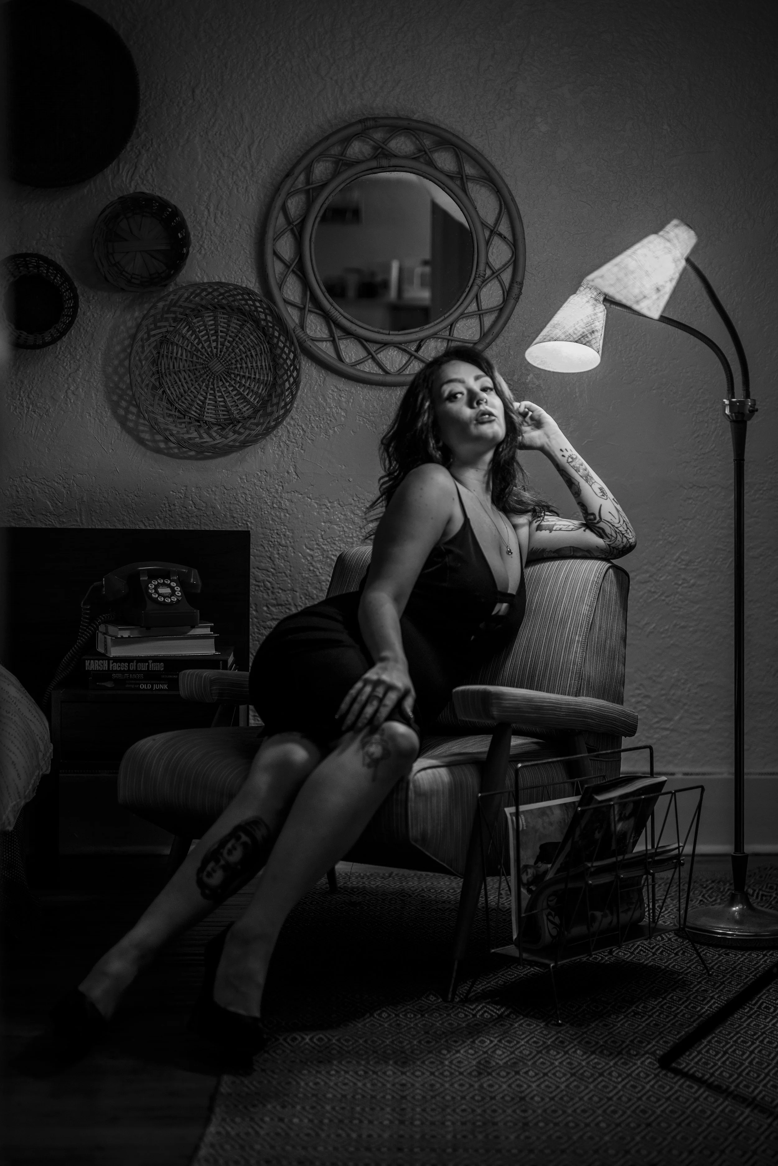 Black and white photo of a woman with tattoos sitting on a vintage armchair in a dimly lit room, surrounded by decorative wall baskets, a mirror, and a floor lamp.