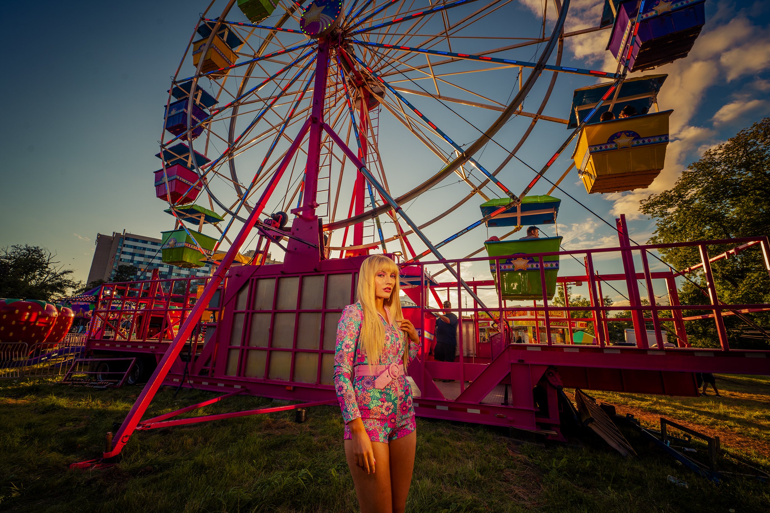 A woman with long blonde hair wearing a colorful floral matching set standing in front of a pink ferris wheel at a fair during sunset.