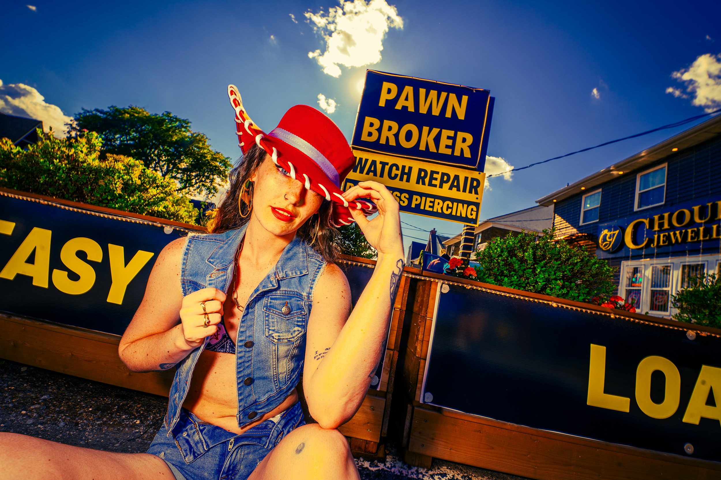 Young woman wearing a red hat, denim vest, and hoop earrings, sitting outdoors at a fair or market, holding her hat and smiling, with signs advertising a pawn broker and watch repair in the background, under a partly cloudy sky.