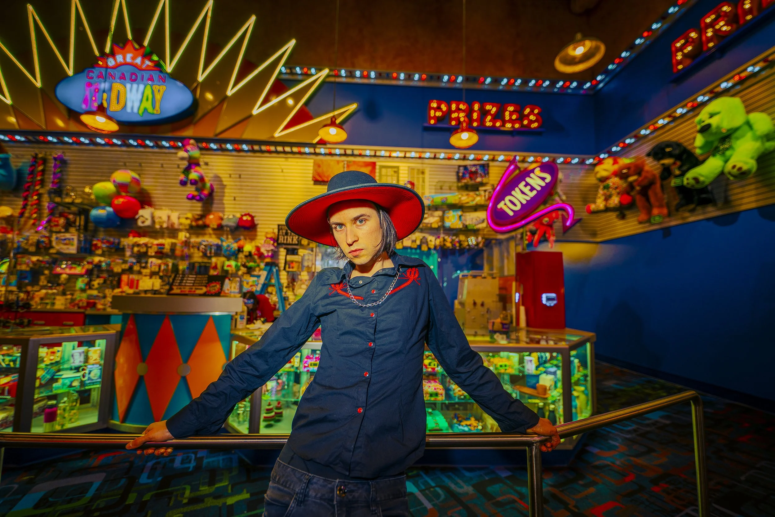 A young person with long dark hair, wearing a large red hat and a dark shirt, standing in an arcade with neon signs that read 'Dream Canadian Holiday,' 'Pizza,' and 'Toys'. The arcade is filled with stuffed animals, bright lights, and colorful toys.