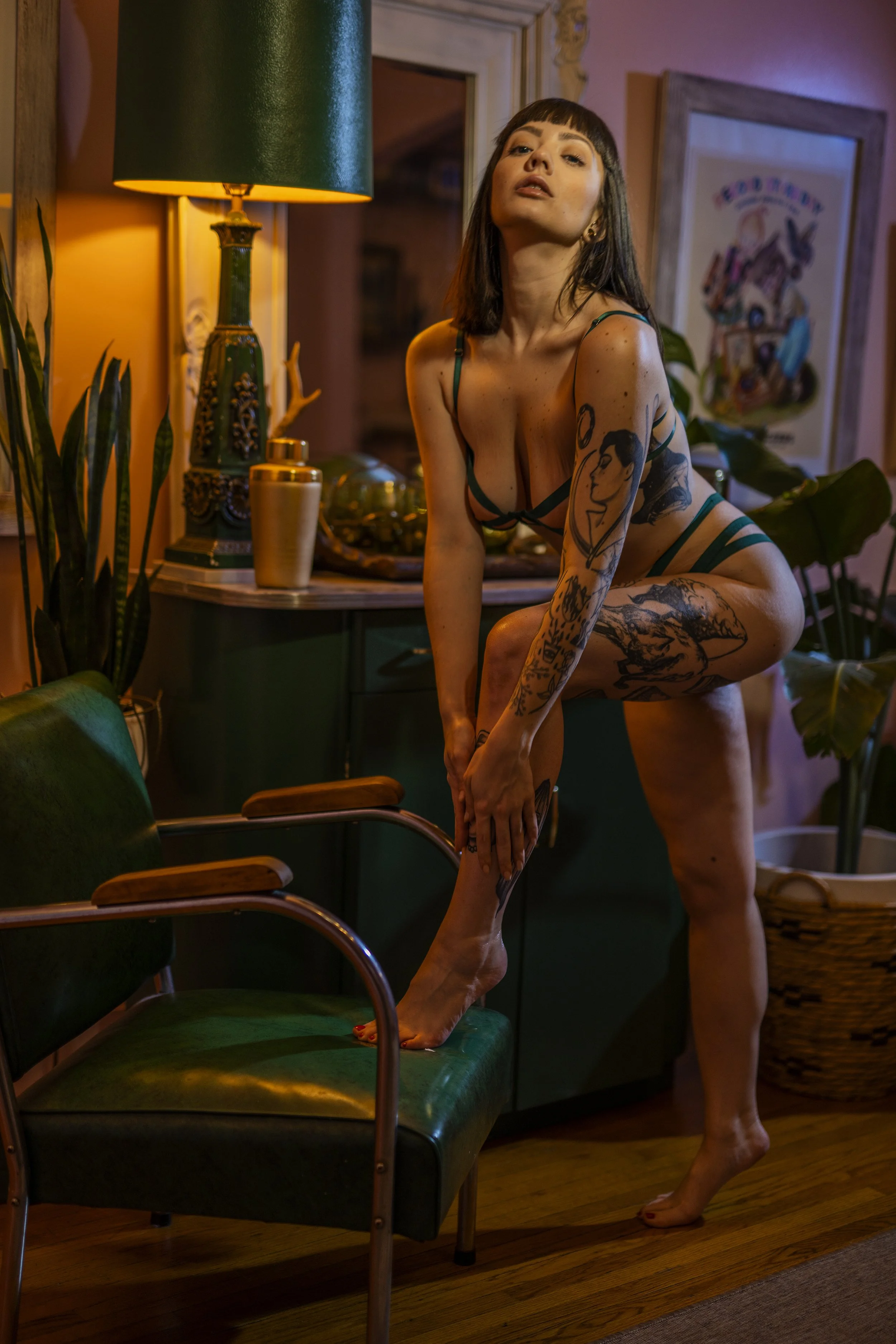 A woman with dark hair and tattoos, wearing lingerie, is standing on one leg with her foot on a green chair. She is holding her other leg with her hands, in a pose in a warmly lit room decorated with plants and framed artwork.