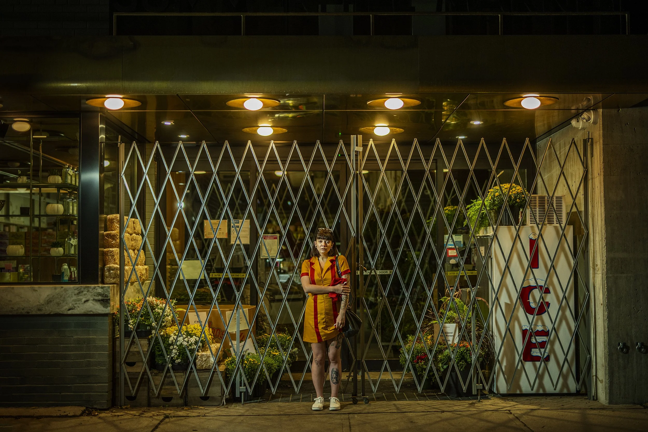A woman standing outside a closed store at night, behind a metal security gate, with flowers and display items inside the store and bright ceiling lights overhead.