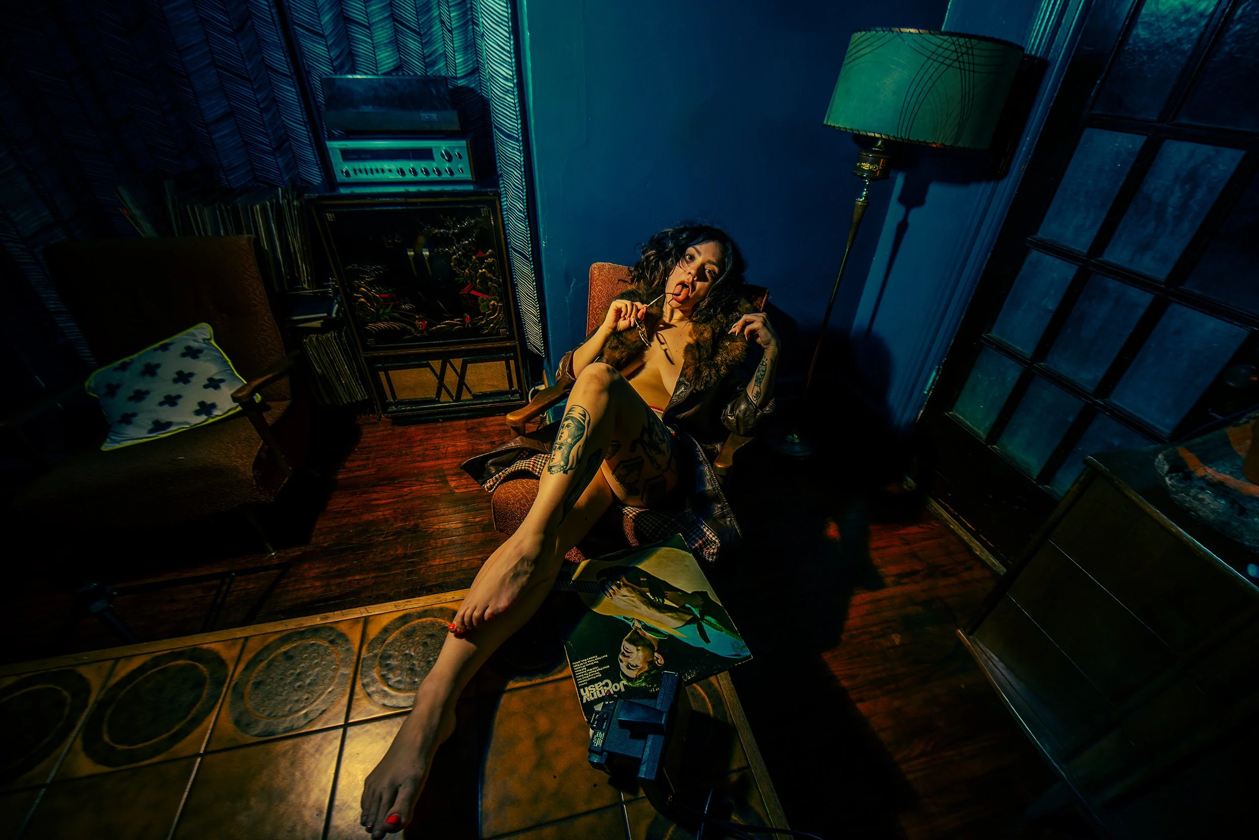 A woman with tattoos, dark curly hair, and bold makeup sitting in a vintage armchair, holding sunglasses in her mouth in a dimly lit room with a lamp, records, and colorful decor.