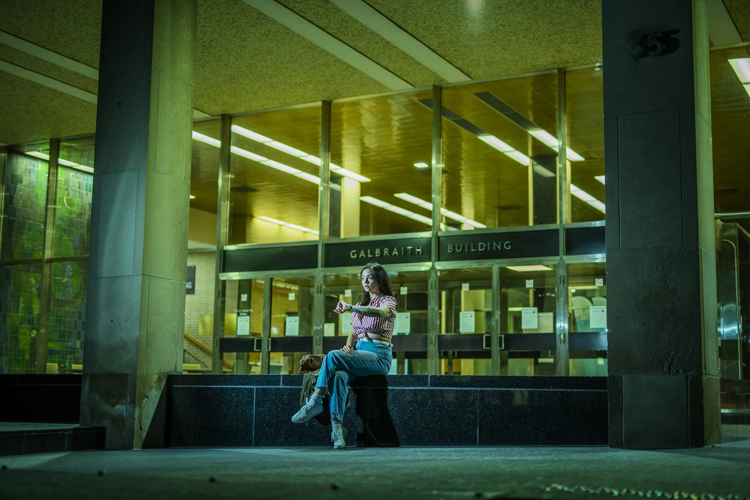 A young woman sitting on a ledge outside the Galbraith Building, looking at her watch in front of a modern, glass-fronted building with illuminated interior lighting.