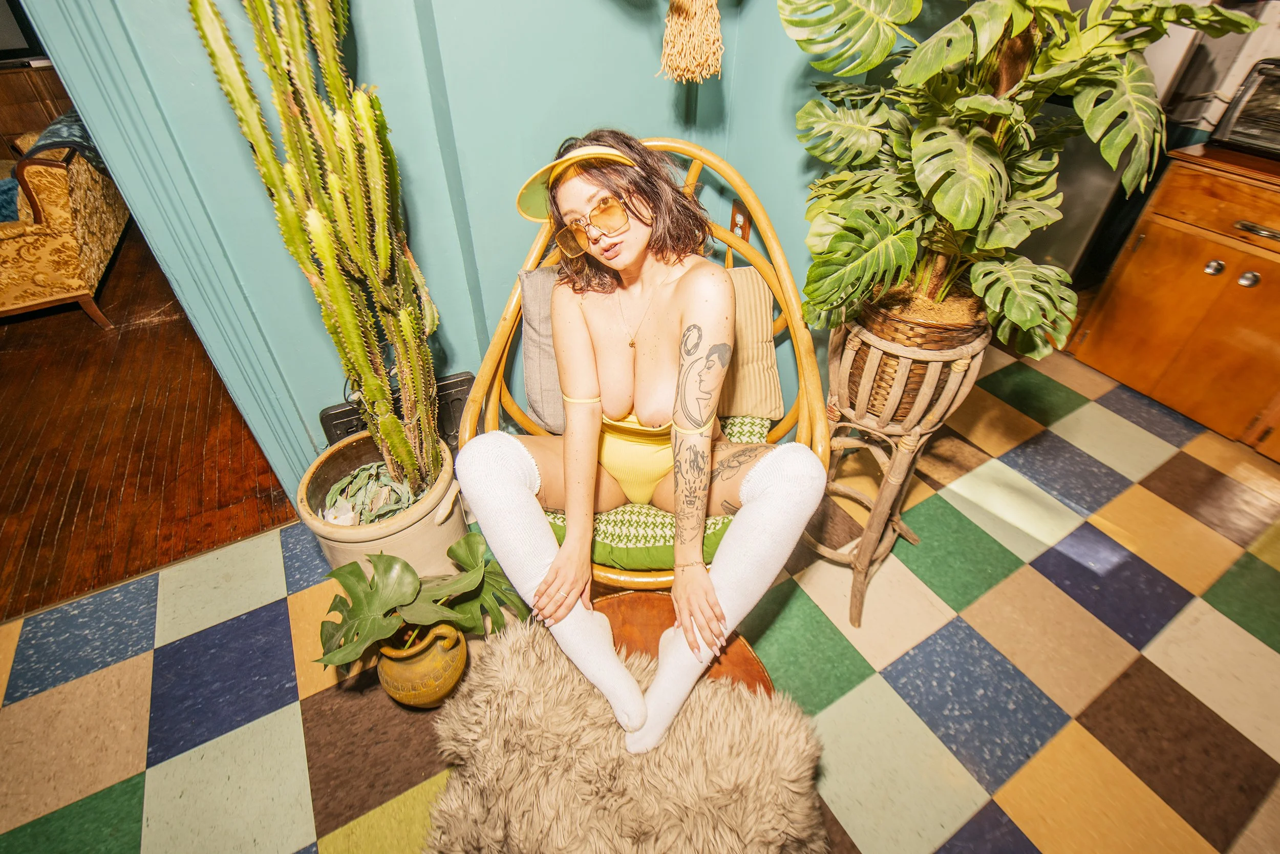 Young woman with tattoos wearing yellow swimsuit and white thigh-high socks sitting cross-legged on a wooden stool surrounded by indoor plants in a colorful room.