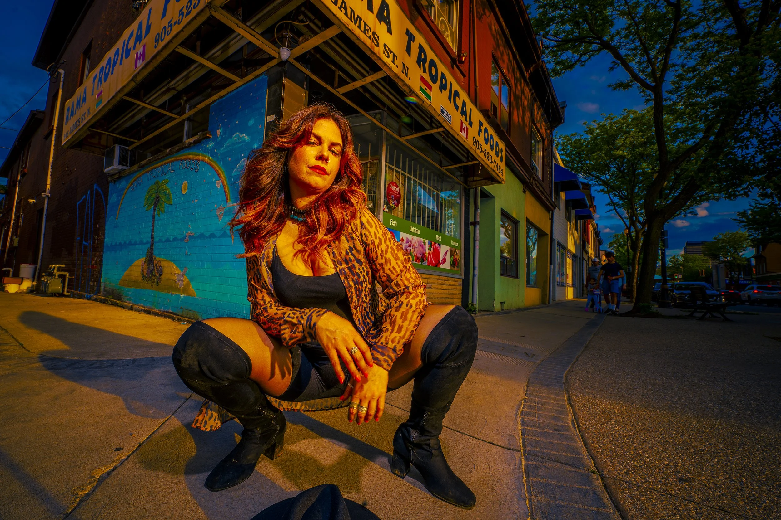 A woman with long red hair and leopard print jacket crouches on the sidewalk at dusk in front of a colorful mural and a storefront with yellow signage. She has a confident expression and is wearing black boots.