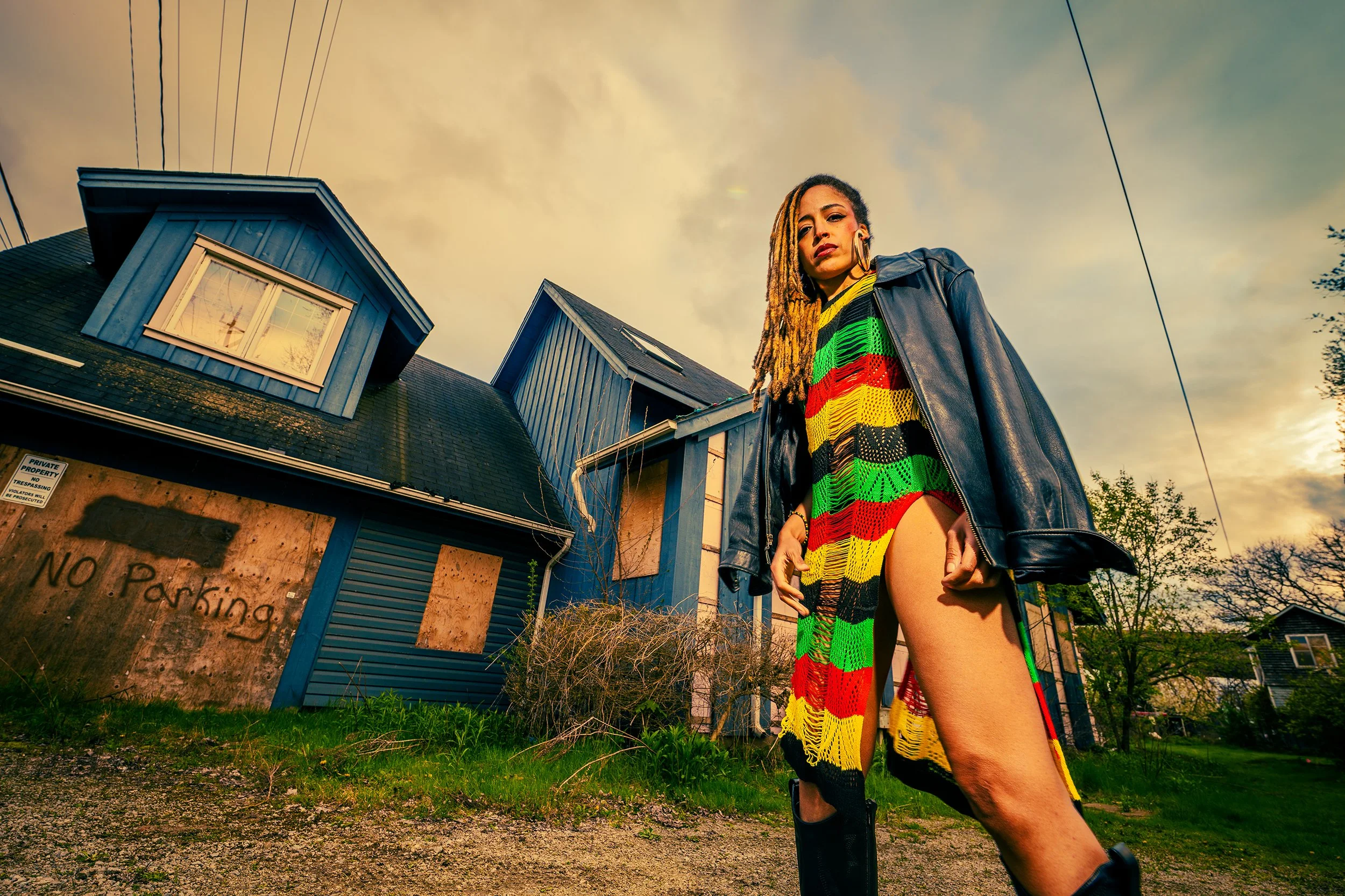 A woman with long dreadlocks, wearing a colorful knitted dress and a black leather jacket, standing outdoors in front of a blue house with boarded-up windows and a sign that says 'No Parking'.