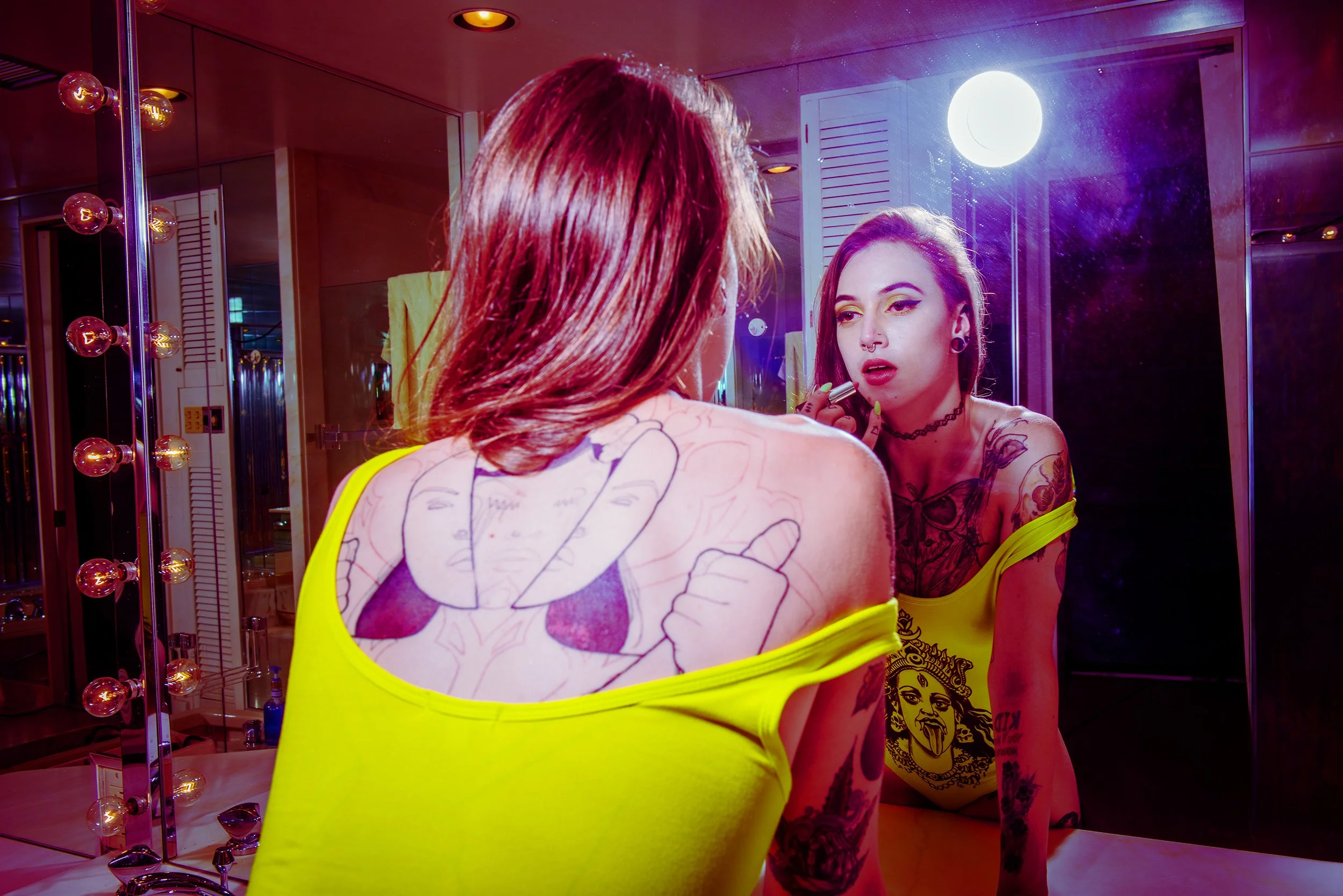 A woman with tattoos on her arms and a choker necklace looks at herself in a mirror while applying lipstick. She wears a bright yellow top with a cartoon graphic on the back. The room has warm lighting with round bulbs and a large white circular ligh