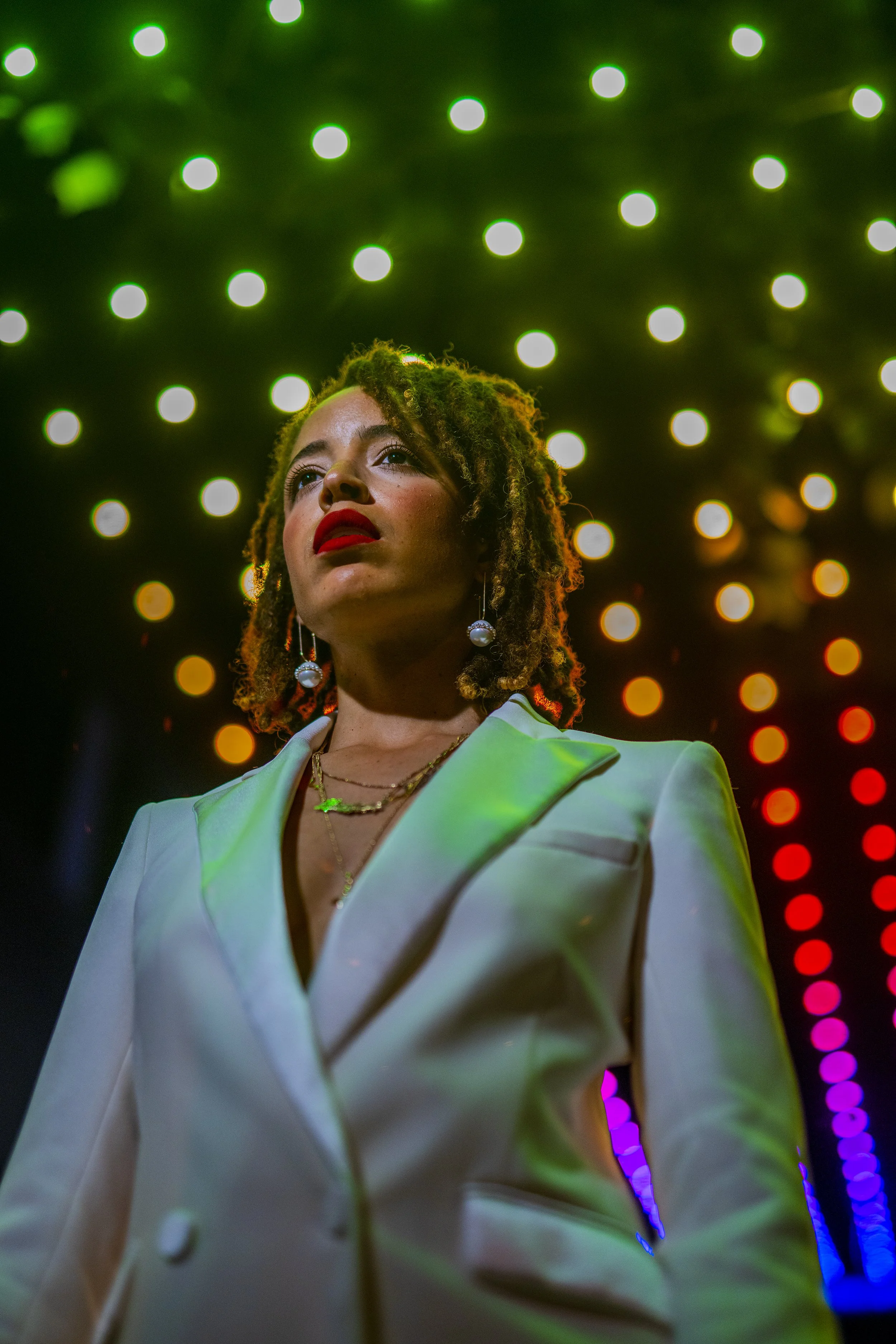 A woman with dreadlocks wearing a white blazer and pearl earrings stands under colorful rainbow lights.