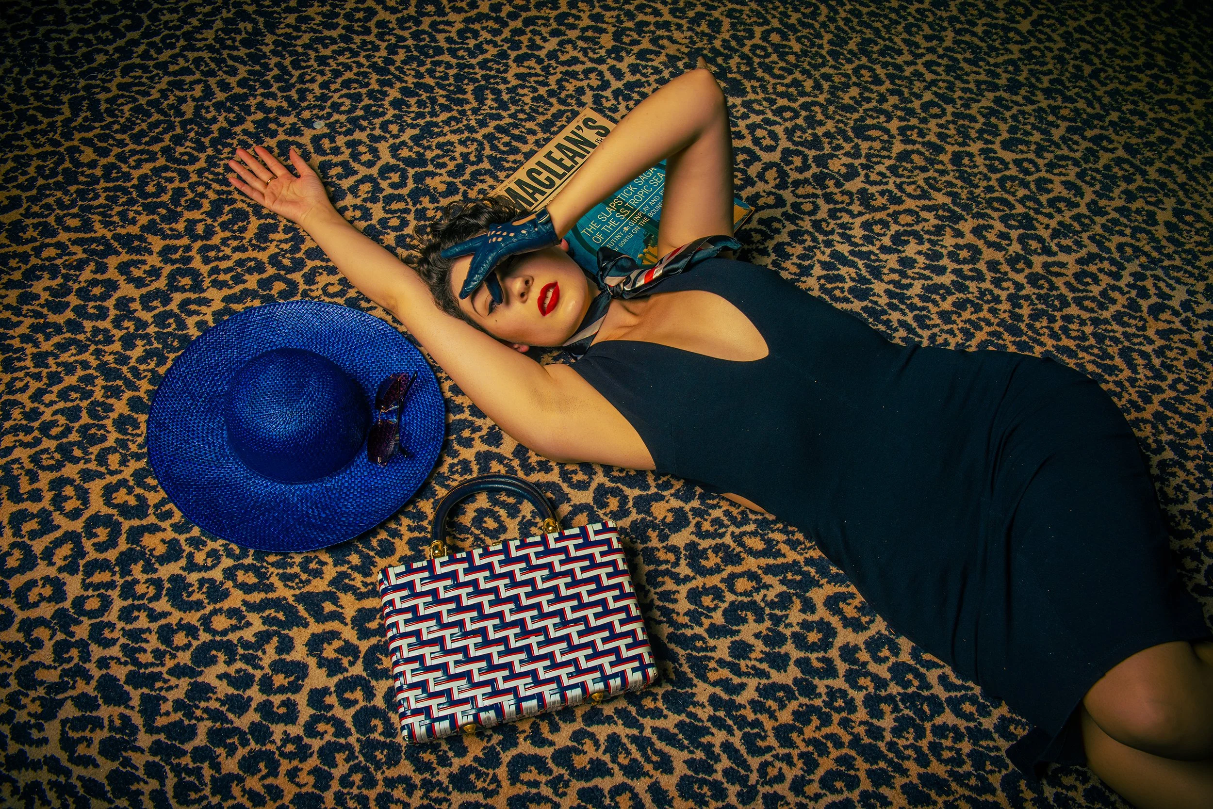 A woman lying on a patterned carpet with a blue wide-brim hat, a patterned handbag, and a magazine titled 'The Slapstick Saga of the Stormy Sea.'
