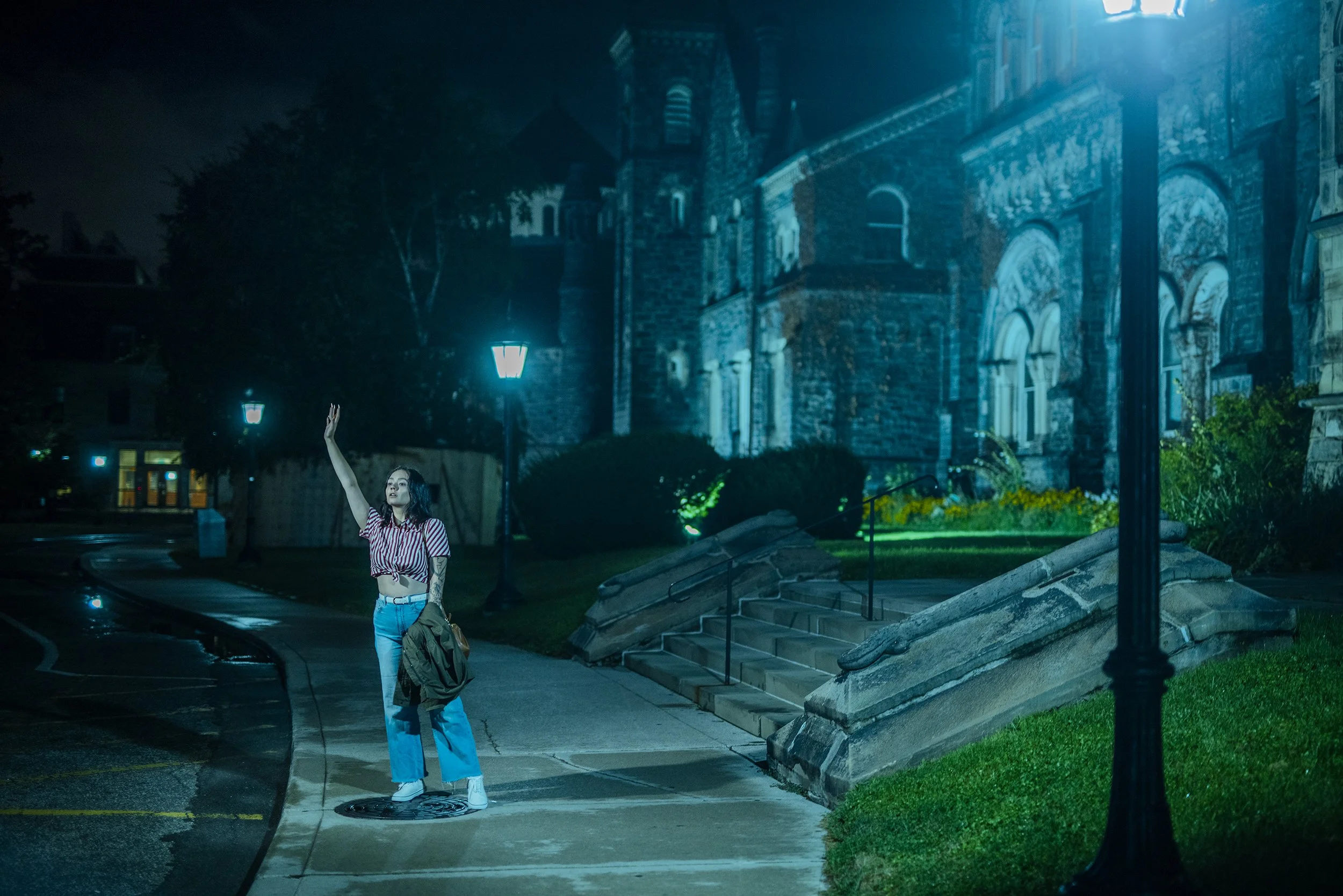 Young woman with backpack waving on sidewalk at night in front of a historic stone building illuminated by streetlights.