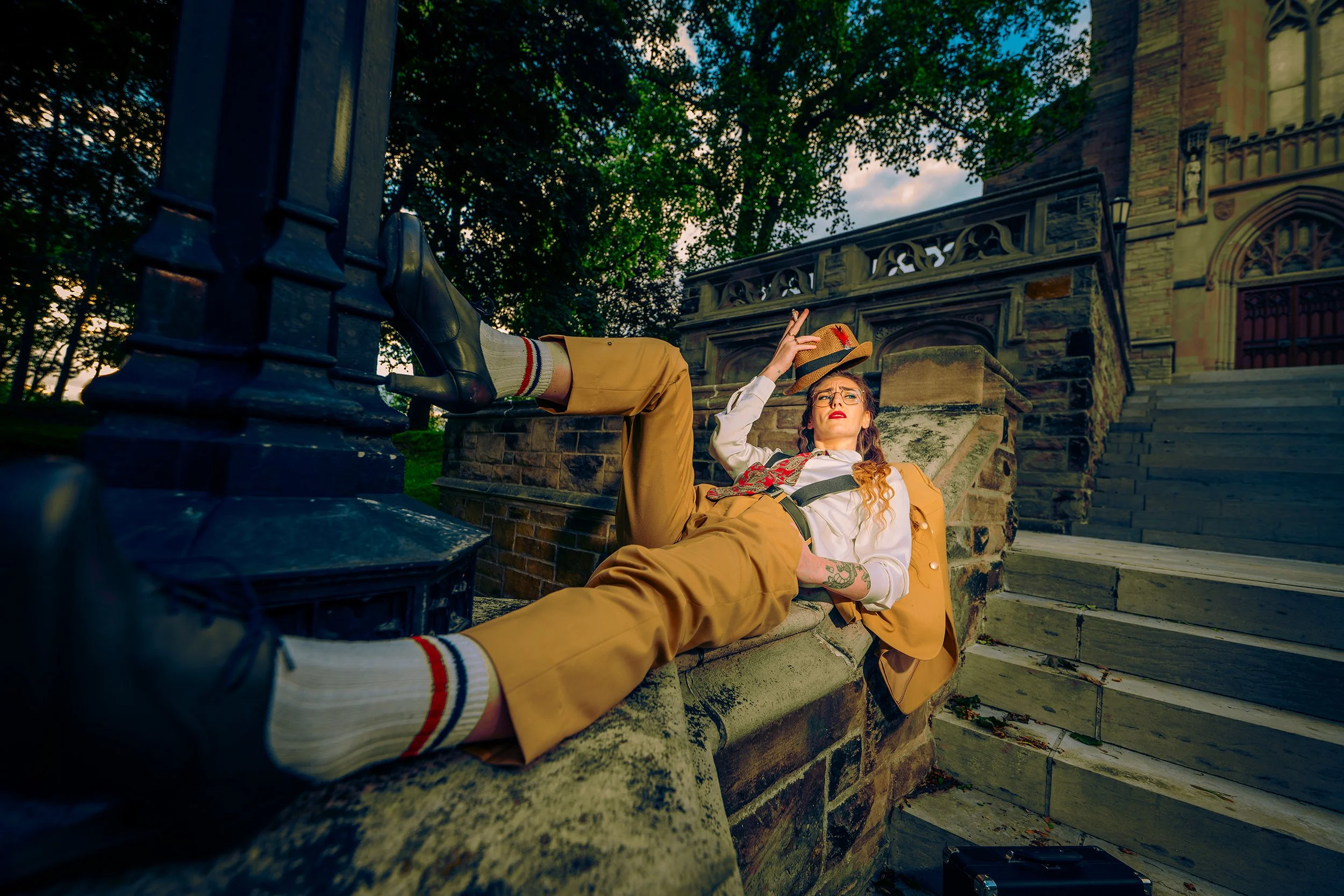 A woman lying on a stone ledge outdoors at sunset, wearing yellow pants, a white shirt, glasses, and a hat, holding another hat in her hand, with a historic building and steps behind her.