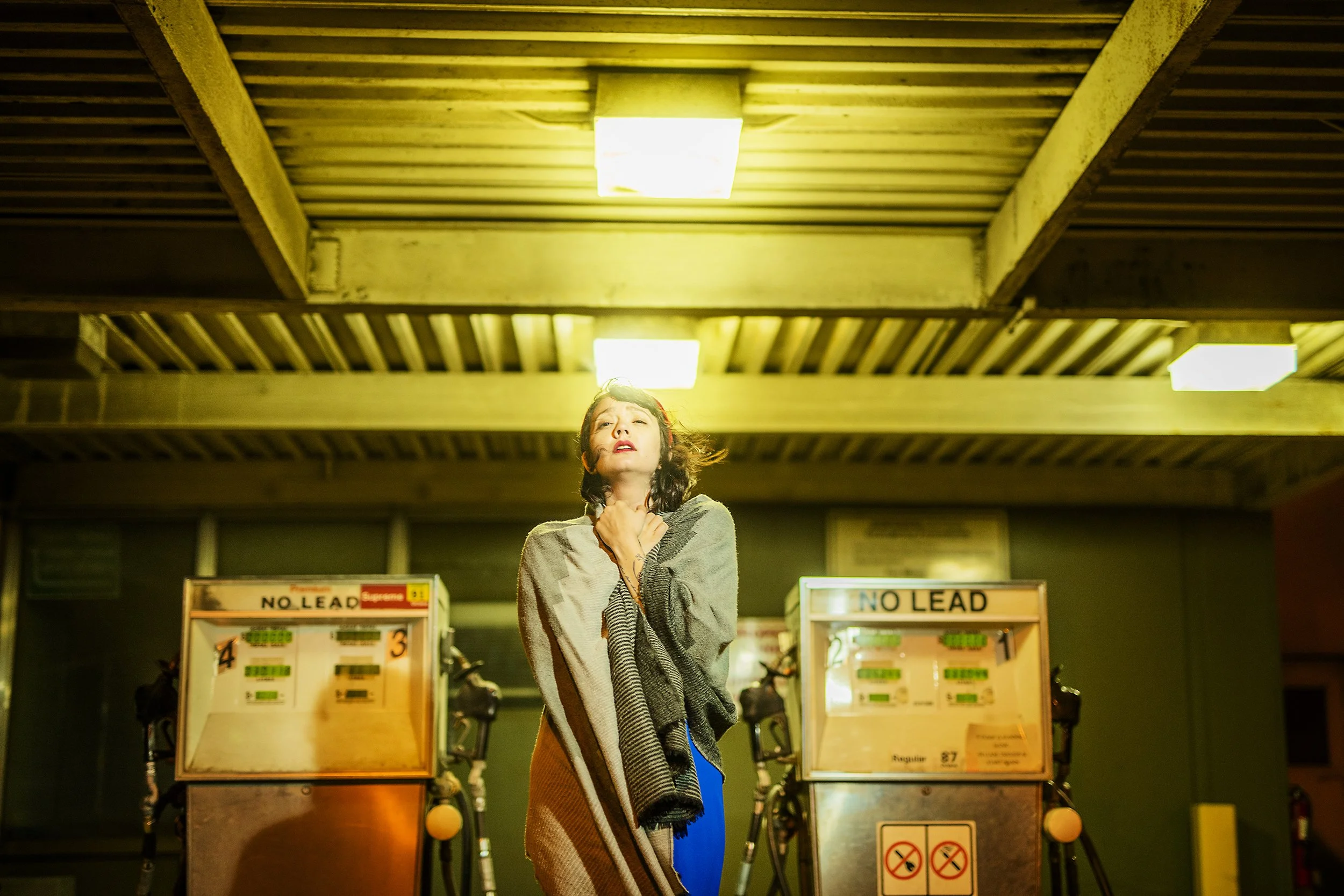 A woman standing under a yellow-lit ceiling near two gas pumps, clutching her chest with an expression of distress.