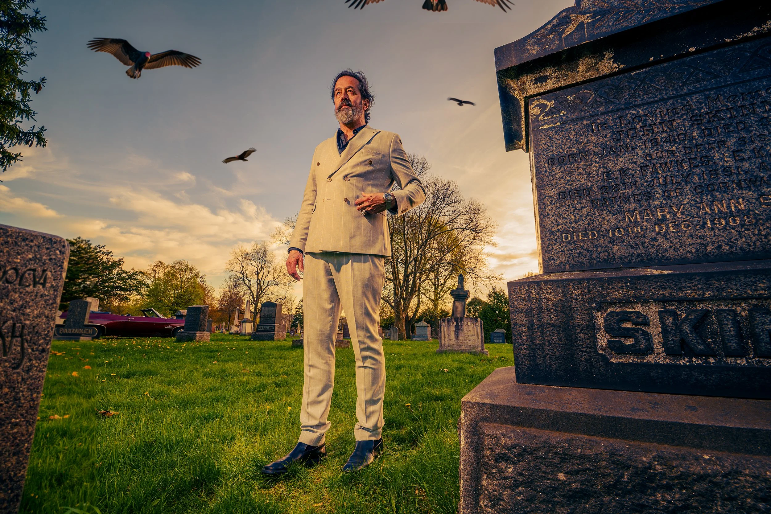 A middle-aged man with dark hair and a beard dressed in a light-colored double-breasted suit standing in a cemetery at sunset. Several birds are flying overhead, and a large stone monument is visible in the foreground to the right, with trees and oth