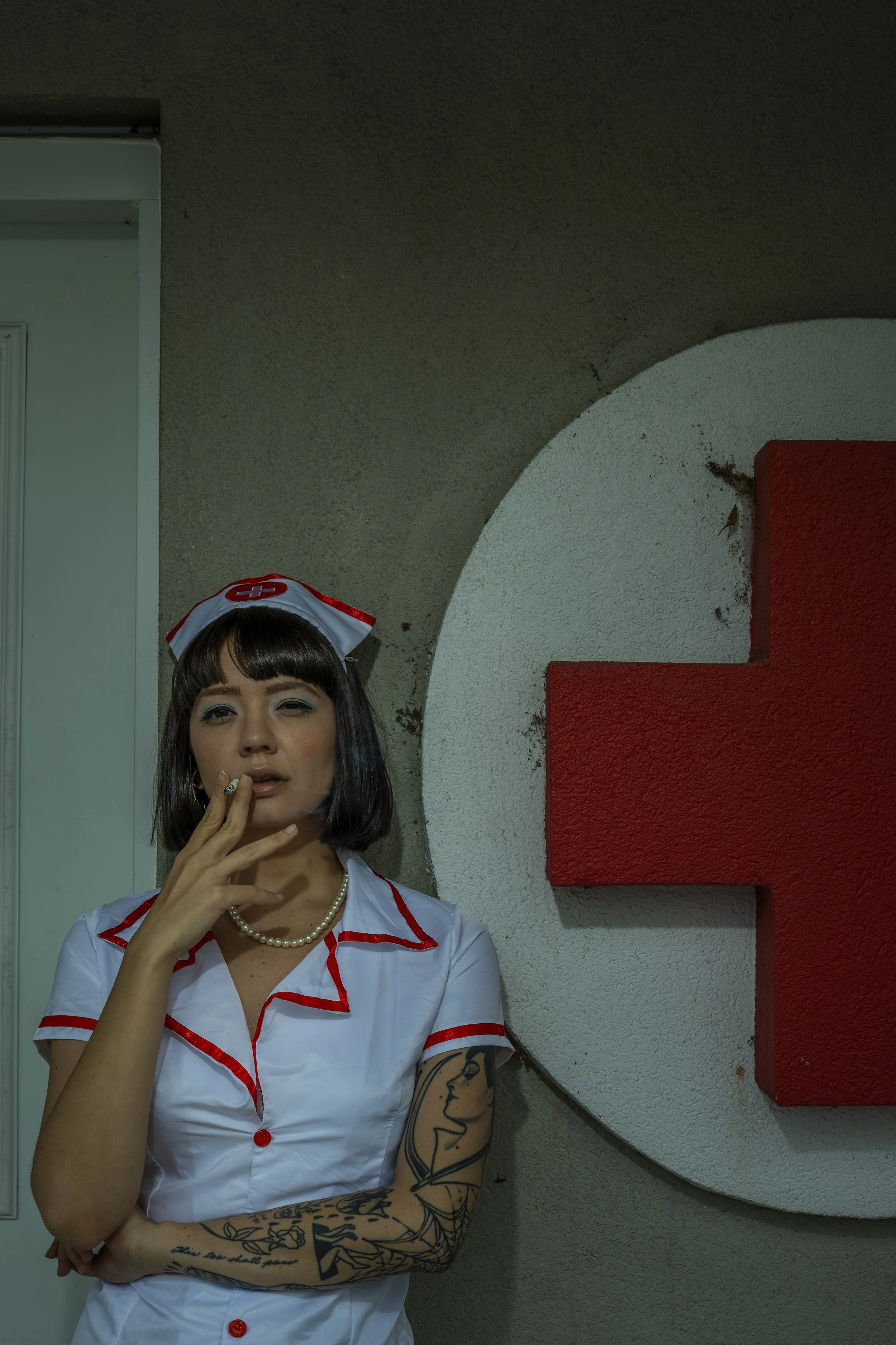 A woman dressed as a nurse with tattoos on her arm, wearing a white outfit with red trim and a nurse cap, standing in front of a wall with a red cross and a round design.