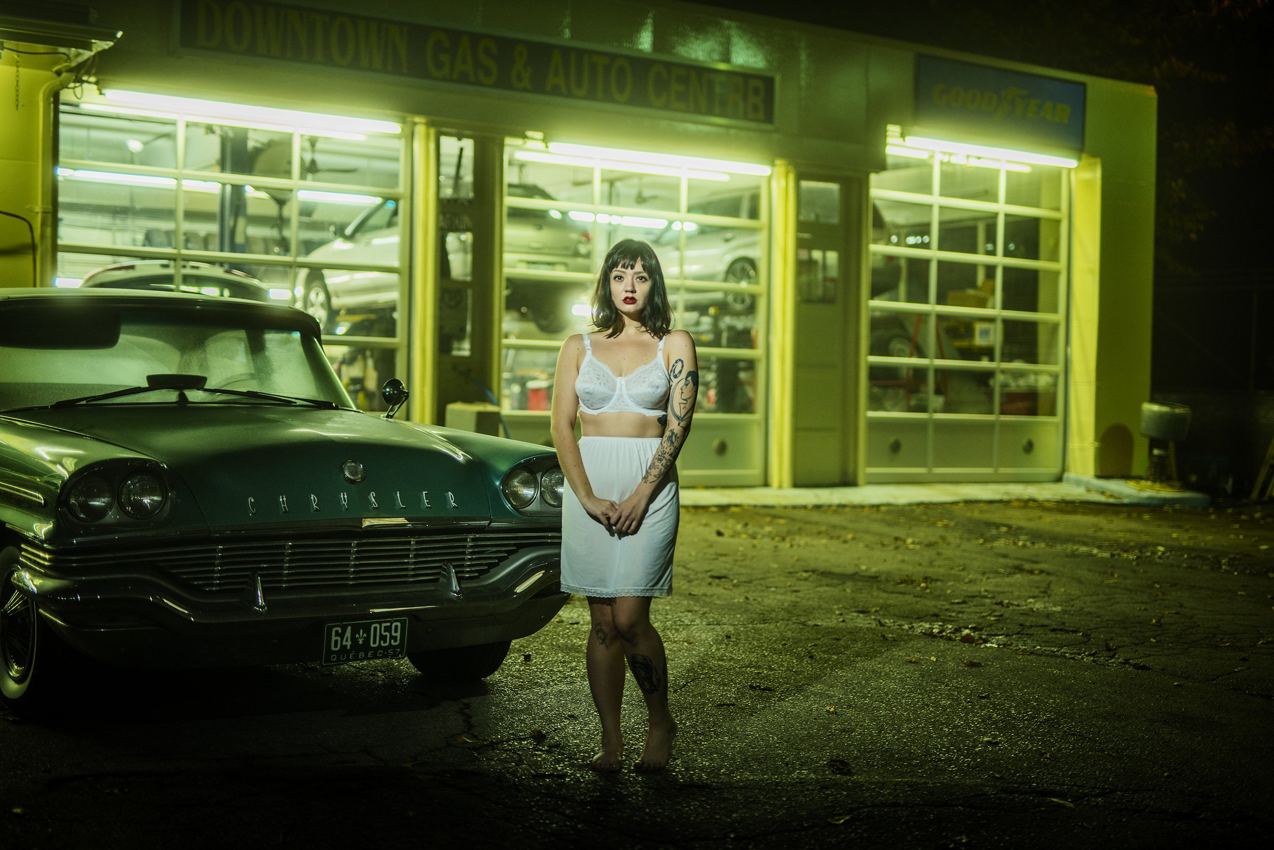 A woman with dark hair and tattoos on her arms stands barefoot outside a yellow garage or auto center at night. She wears a white lace bra and a white skirt. A classic black Chrysler car is parked nearby, and the garage's glass windows reveal multipl