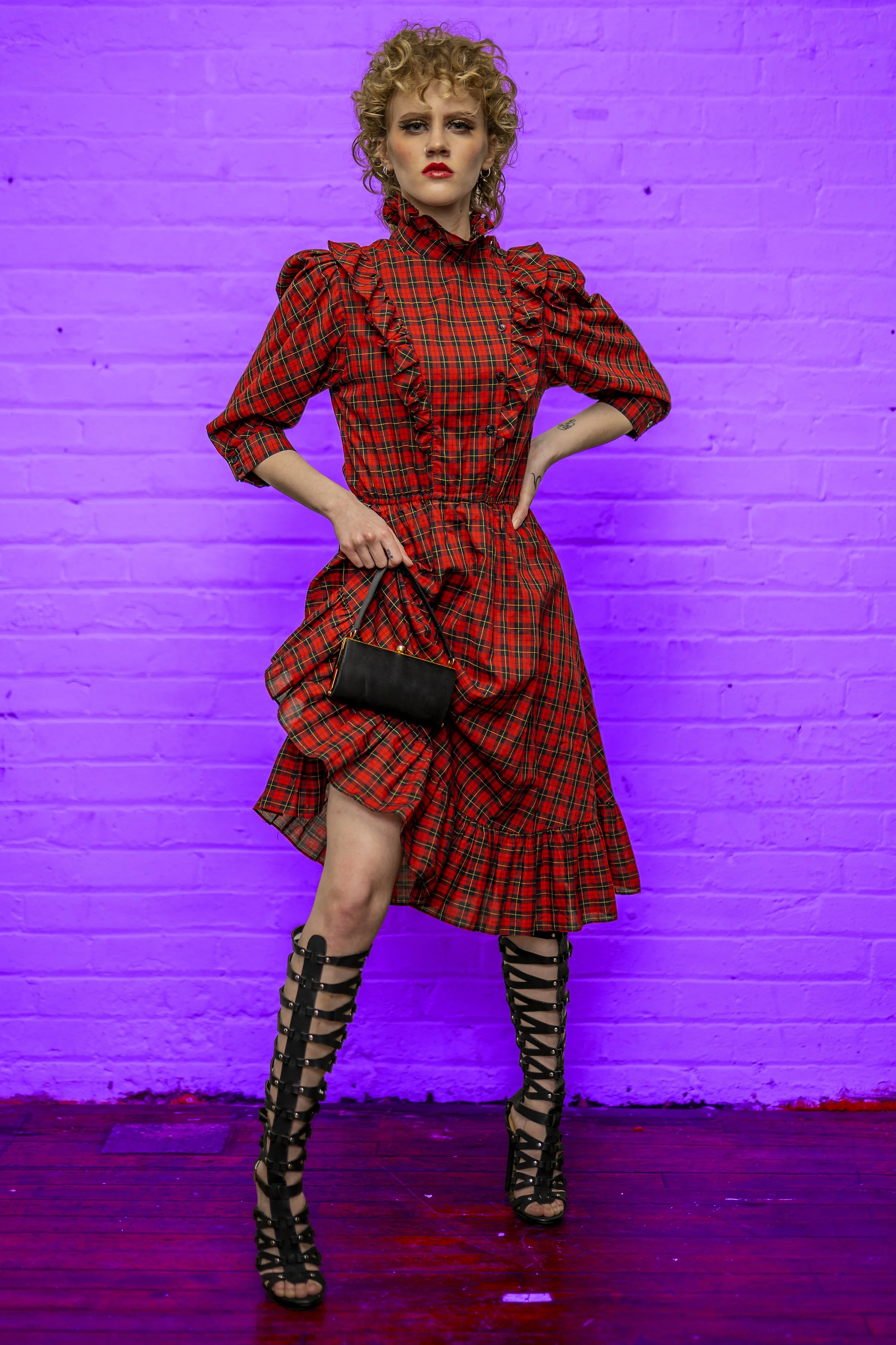 A woman with curly blonde hair wearing a red plaid dress with ruffles, holding a small black handbag, standing against a purple-lit brick wall, wearing black gladiator sandals.