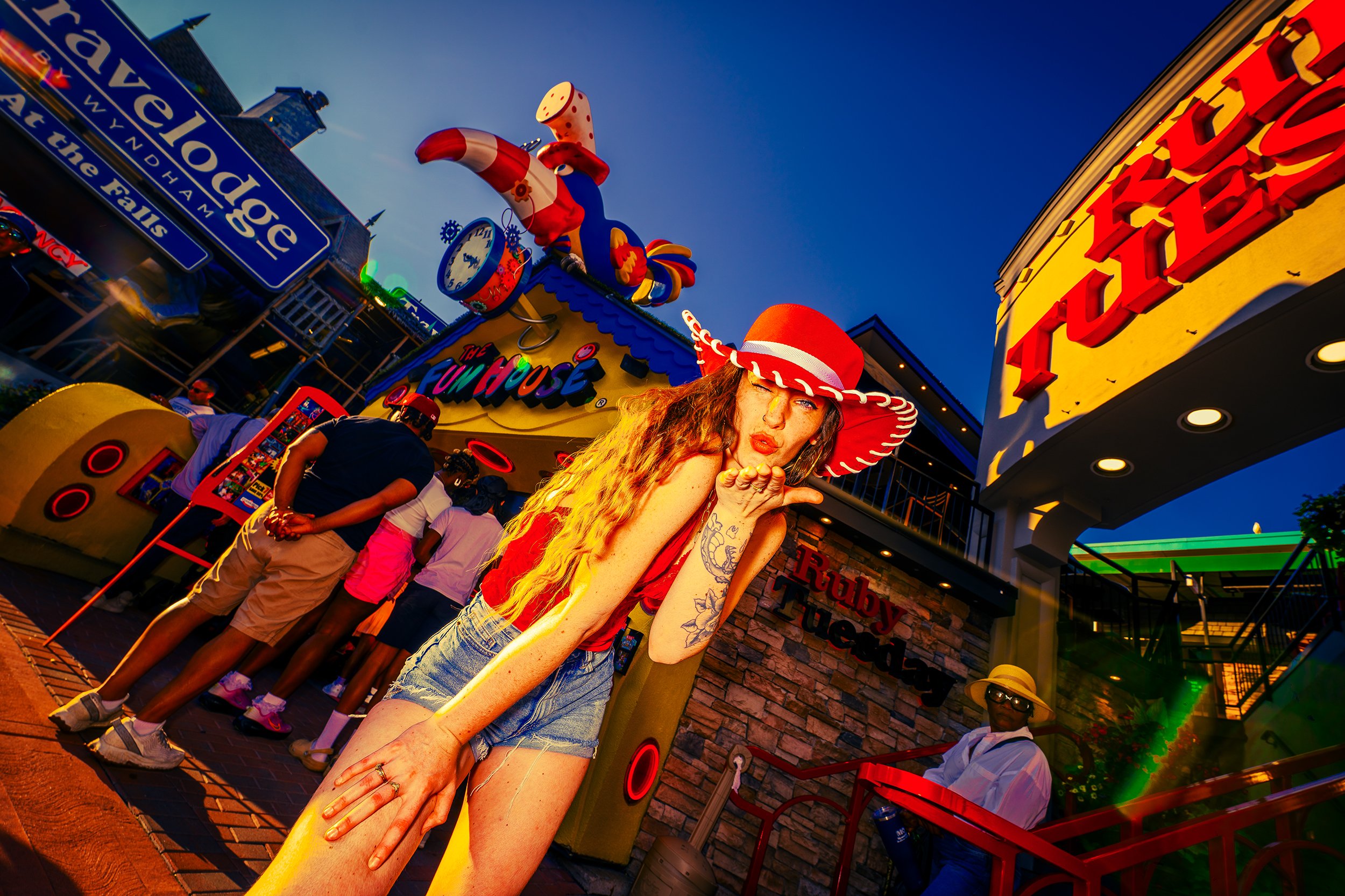Having fun at an amusement park with a woman wearing a large red hat, blowing a kiss, and standing behind a yellow pole. Other visitors are in line, and colorful signs and a large cartoon rooster are visible.