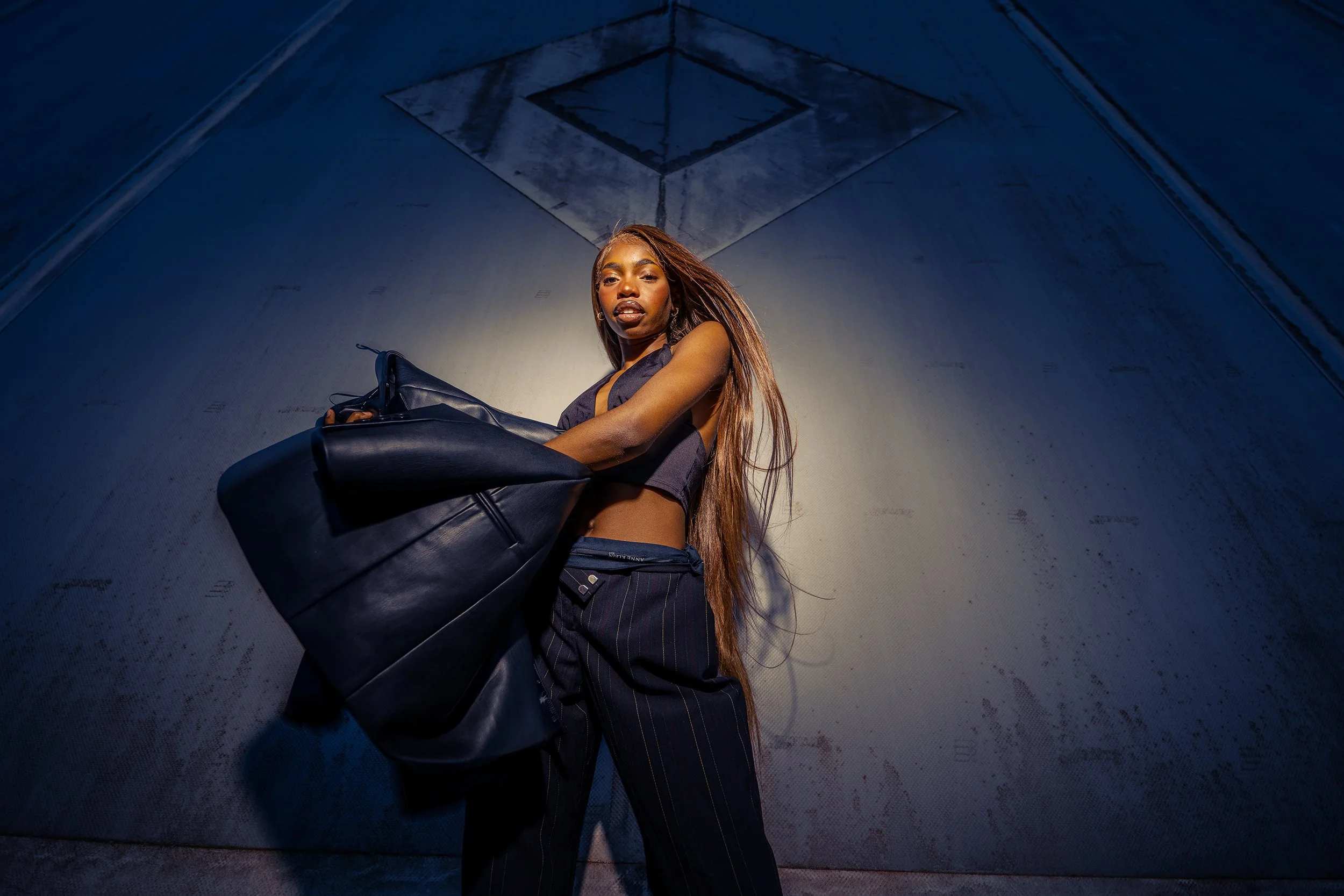 A young woman with long hair, dressed in a black crop top and pinstriped pants, is standing in a dimly lit, industrial-looking tunnel, holding a large black leather bag, looking at the camera.