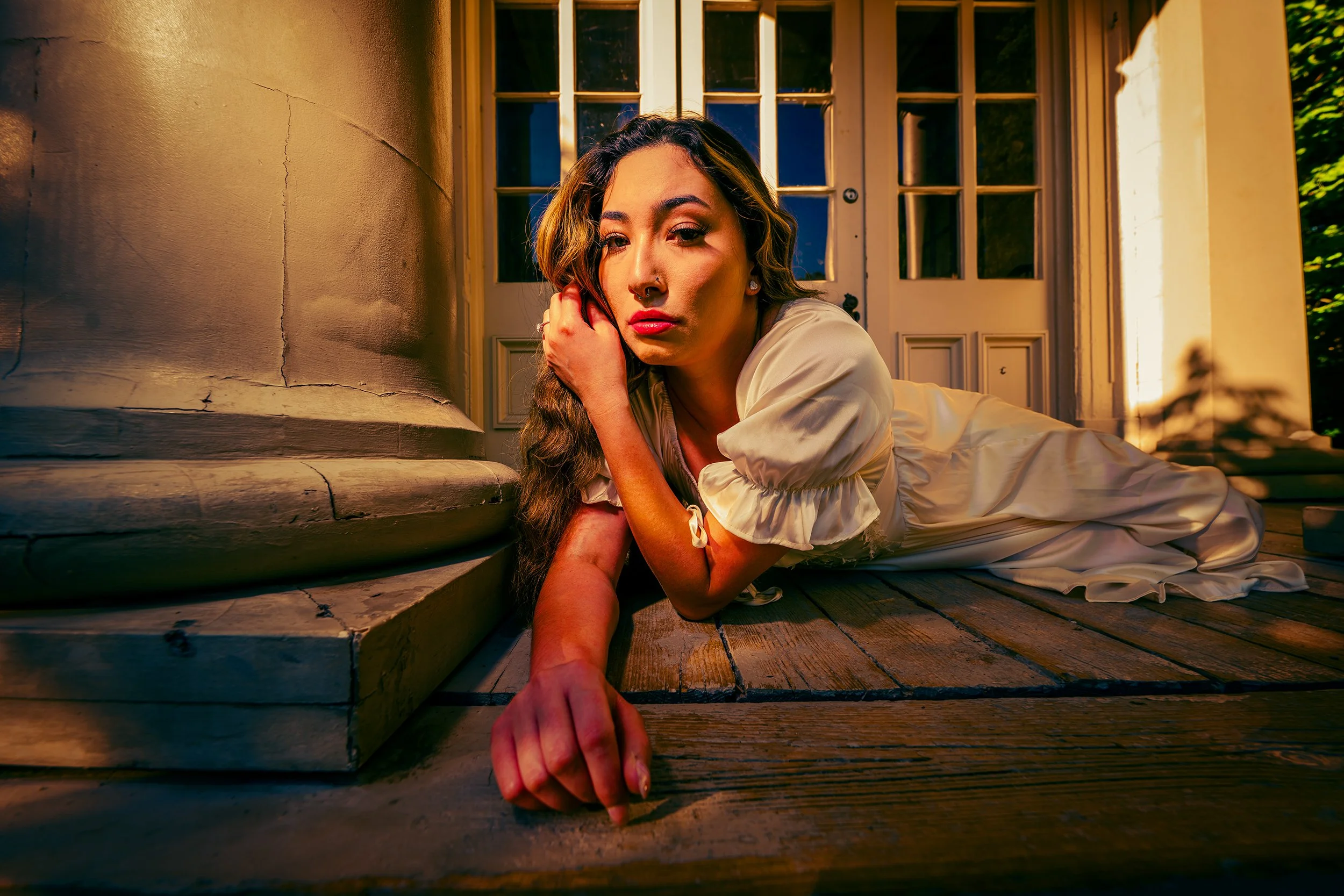 A young woman with long wavy hair and a nose piercing lies on a wooden porch, resting her head on her hand, in front of a door with multiple glass panes. The setting is illuminated by warm sunlight, creating a cozy atmosphere.