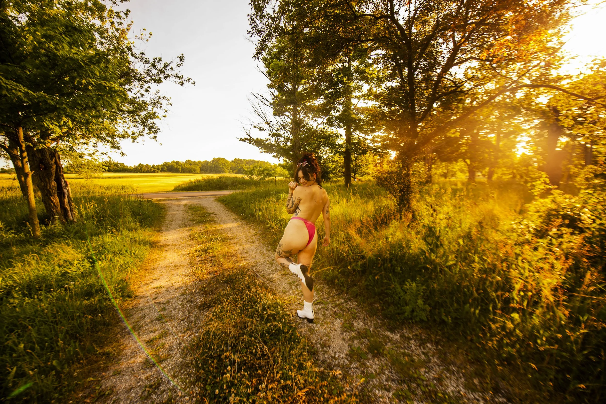 A woman with tattoos on her arm and leg, wearing pink shorts, white socks, and sneakers, stands on a dirt path in a grassy field at sunset, turning and looking back at the camera.