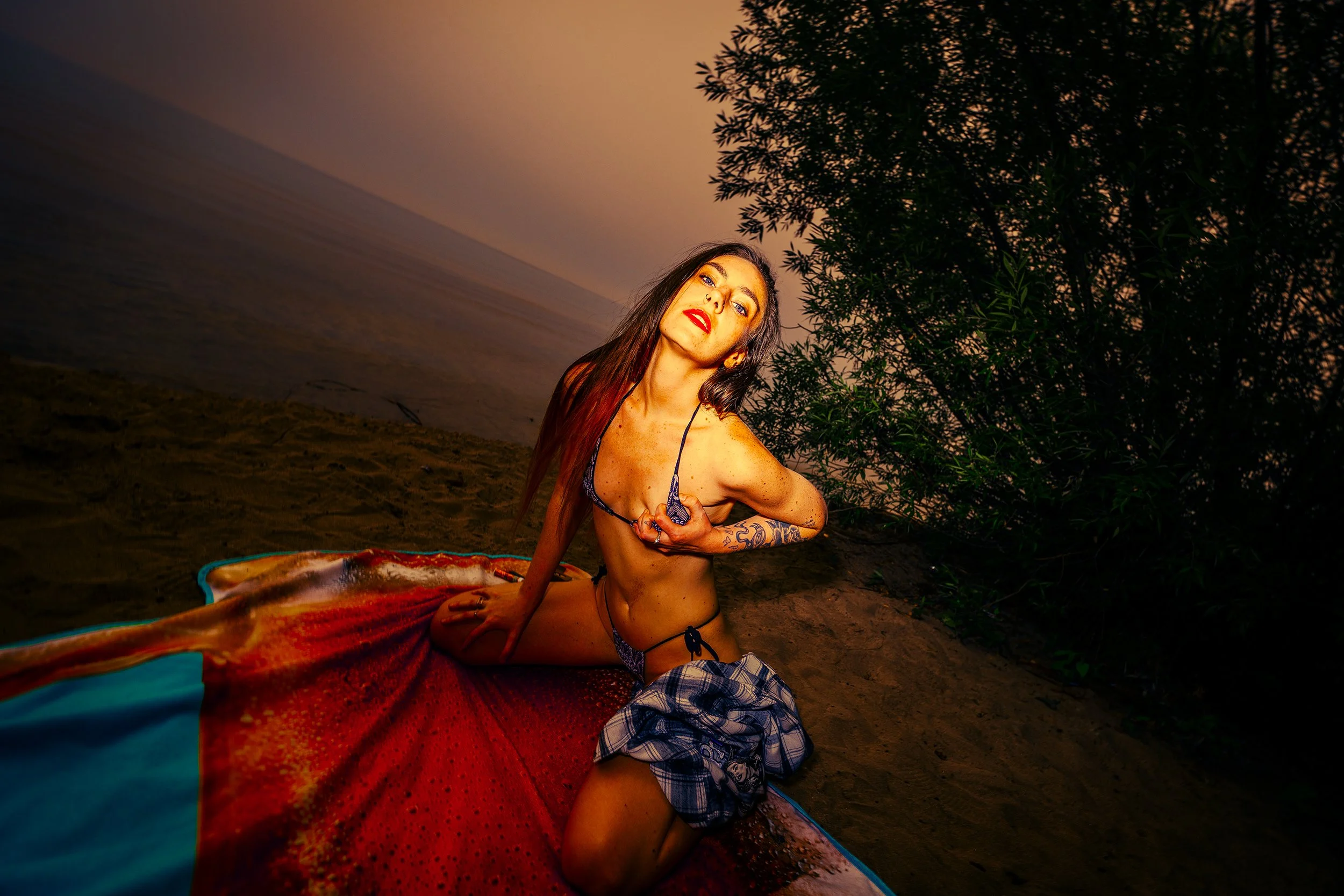 A young woman with long dark hair, wearing red lipstick, a black bikini top, and plaid shorts, is kneeling on a sandy beach at sunset. She is looking at the camera with a confident expression, holding her bikini top with one hand. There are trees to 