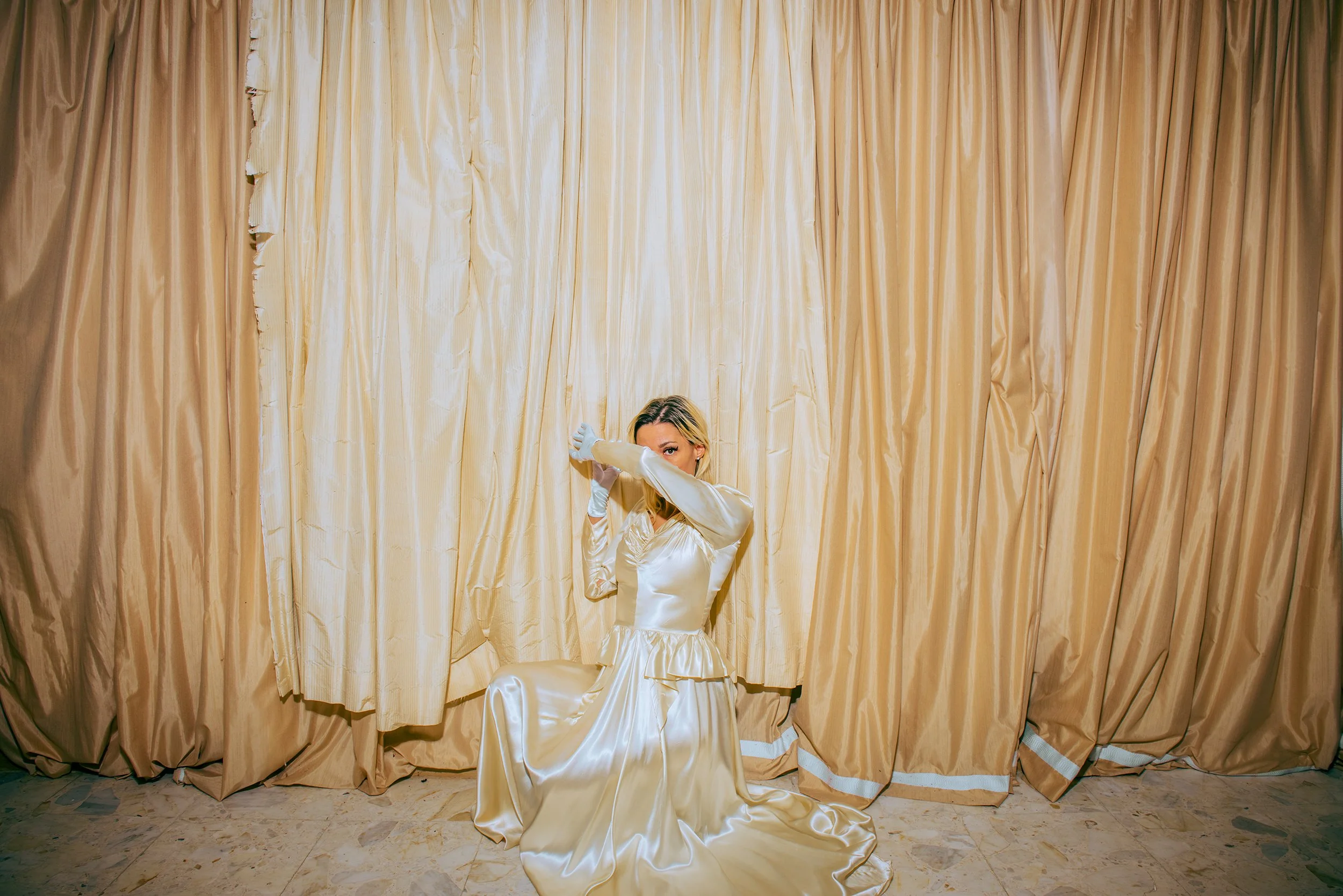 A woman in a long, shiny cream-colored satin dress and white gloves is kneeling on the floor in front of golden curtains, with her right arm covering her face.
