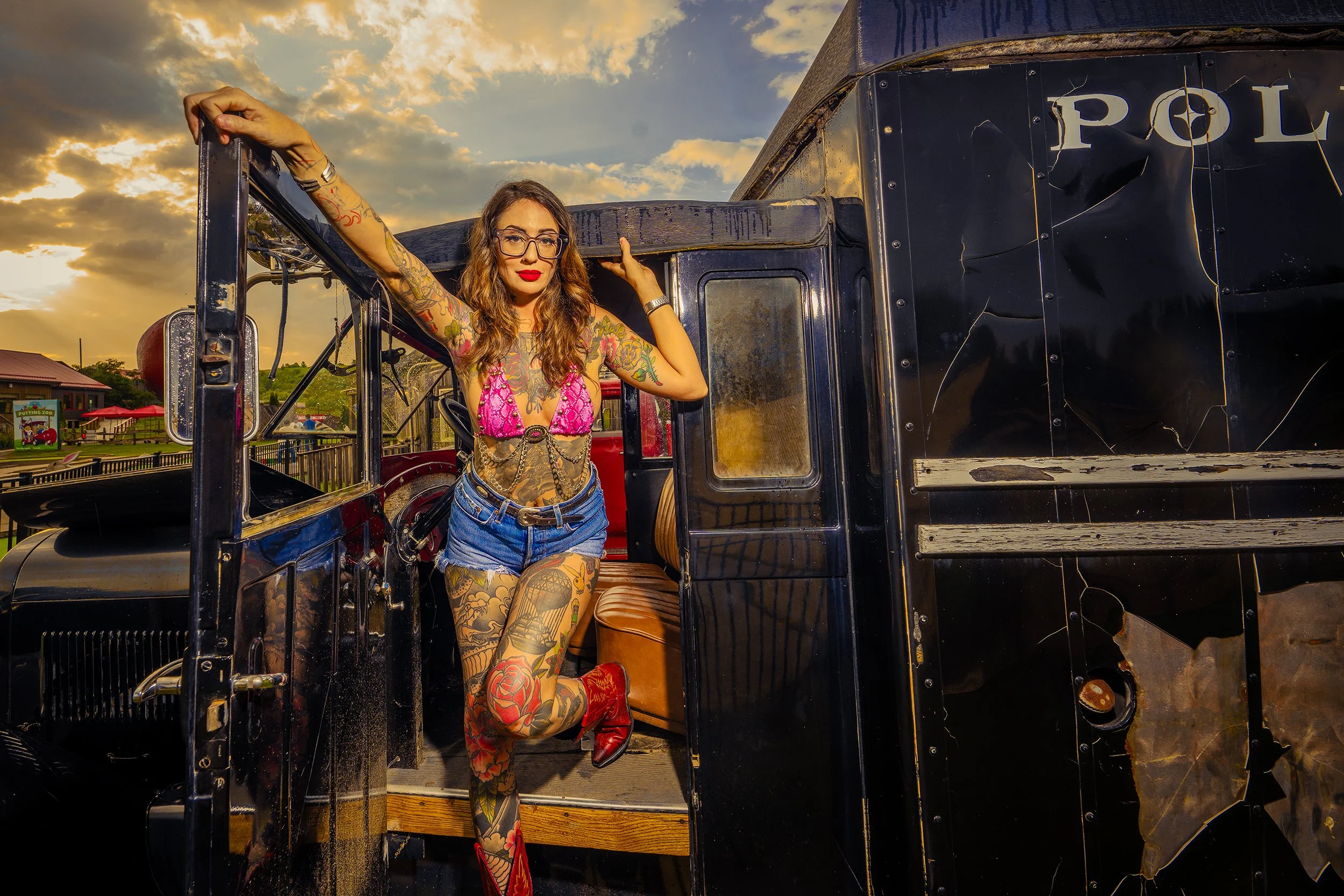 A woman with tattoos, wearing glasses, a pink top, denim shorts, and red boots, leaning on the open door of a black police truck during sunset.