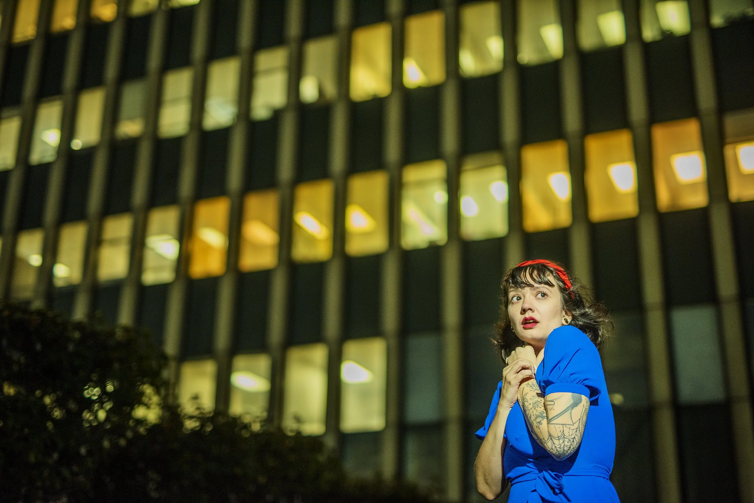 A woman with tattoos, wearing a blue top, stands outdoors at night in front of a building with lit windows, her hands clasped to her chest, looking up.