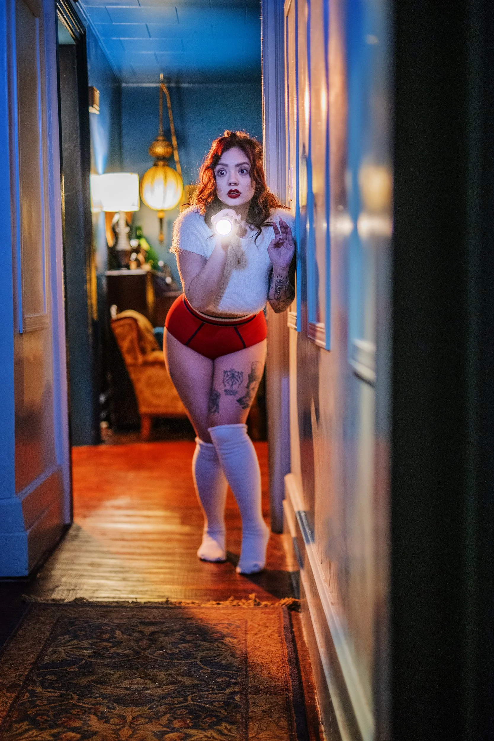 A woman with red hair, tattoos, and wearing red shorts, a white top, and white thigh-high socks, holding a flashlight and looking surprised in a dimly lit hallway with warm lighting and vintage decor.