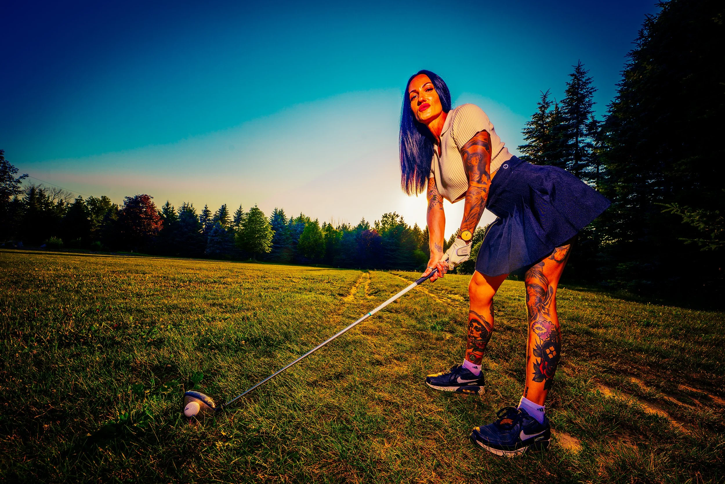 A woman with long blue hair and tattoos, wearing a beige crop top, dark shorts, and sneakers, playing golf outdoors during sunset.