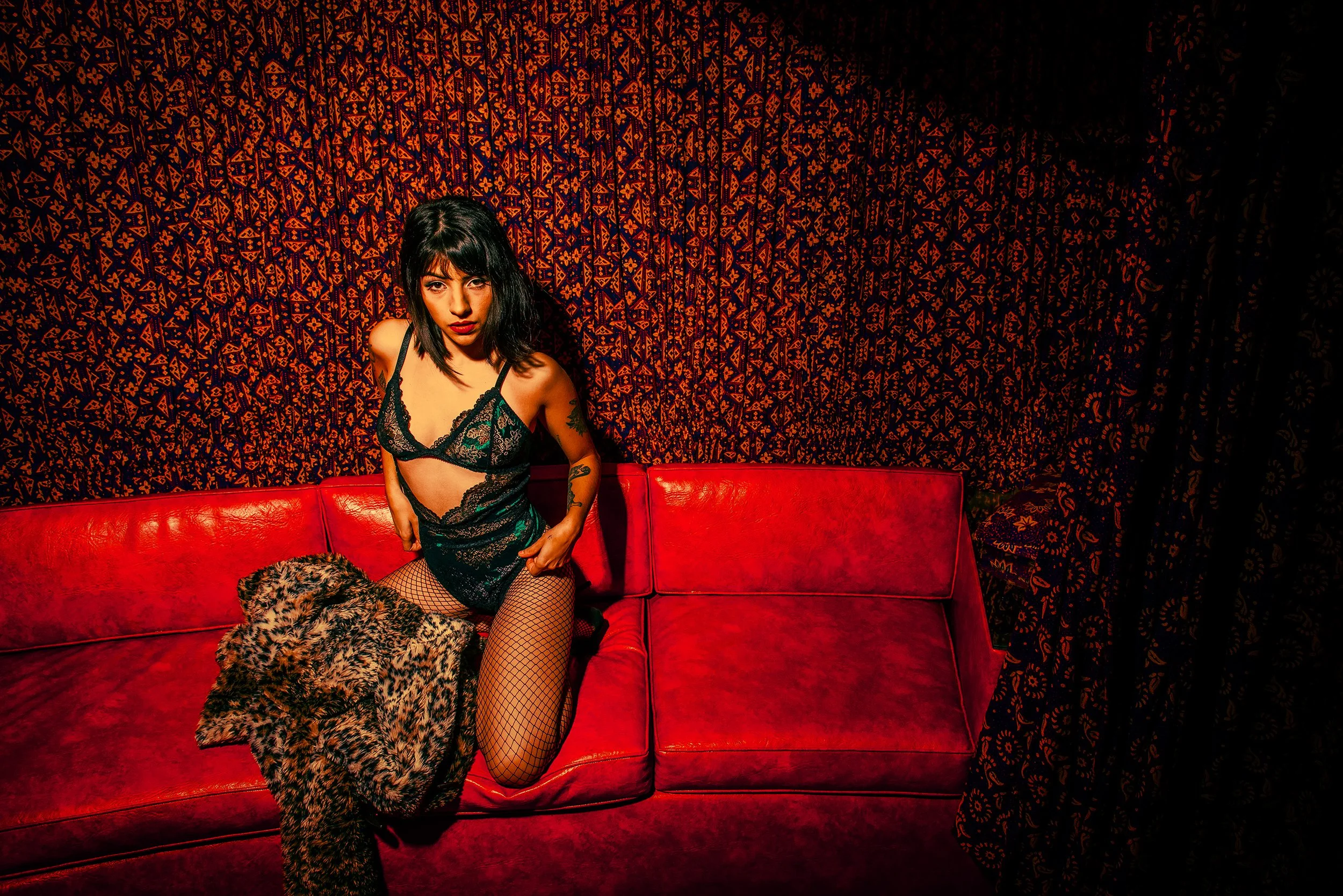A woman with black hair and tattoos on her arm, dressed in black lace clothing, sitting on a red couch in a room with dark red and black patterned wallpaper, looking at the camera.