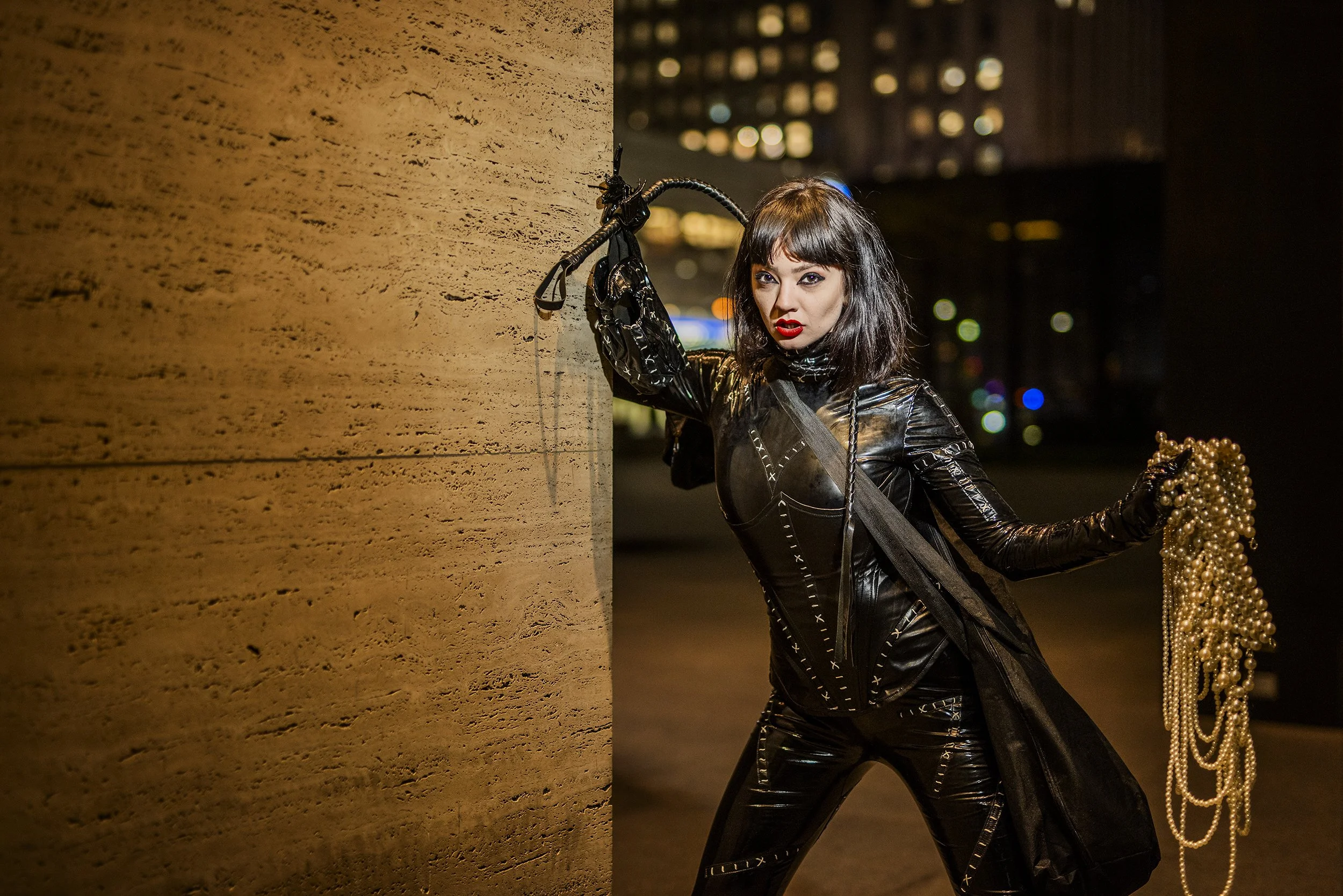 A woman with dark hair and red lipstick dressed in a shiny black leather outfit, holding pearl necklaces and standing next to a beige stone wall at night with city lights in the background.