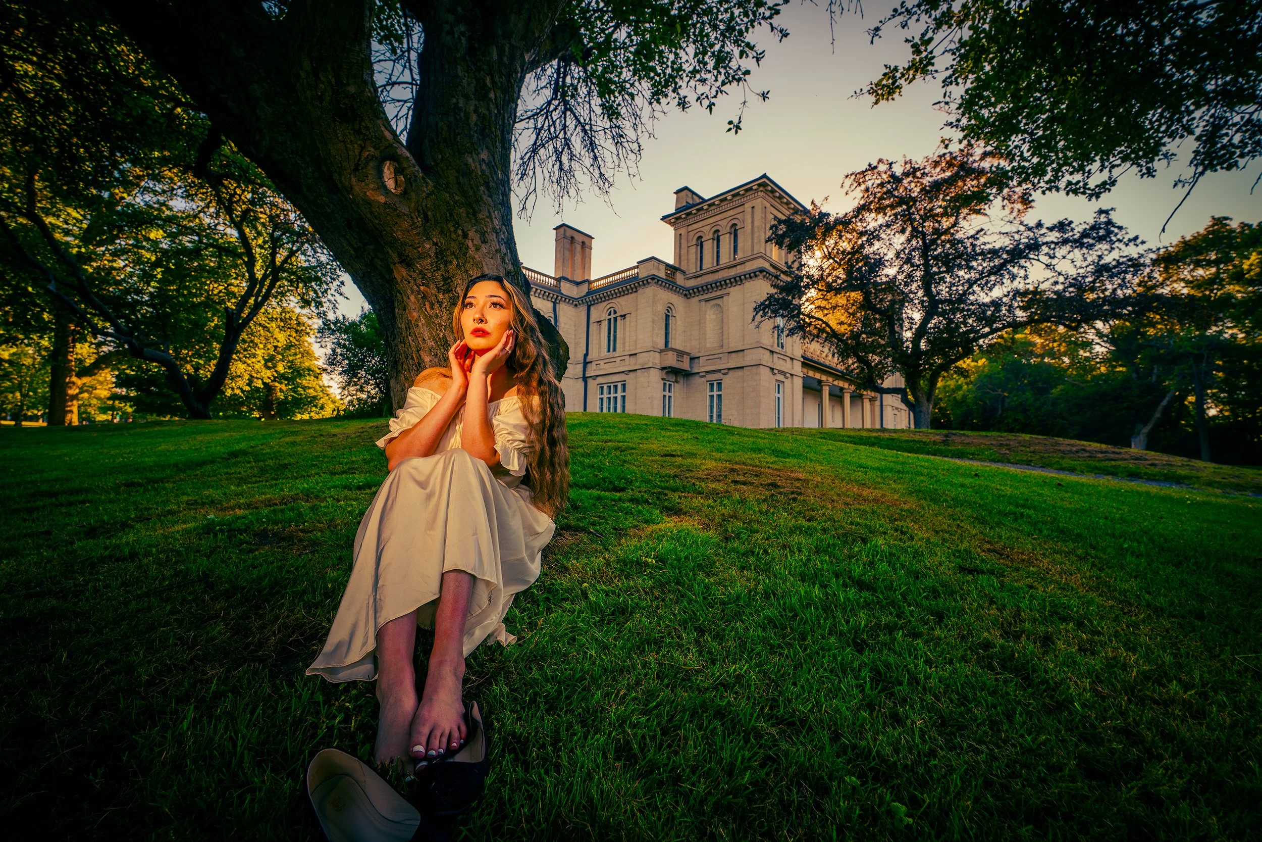A woman with long wavy hair, sitting on a grassy lawn near a large tree, with a historic building in the background. She is wearing a light-colored off-shoulder dress and looks thoughtful, with her hands touching her face. The scene is during the lat