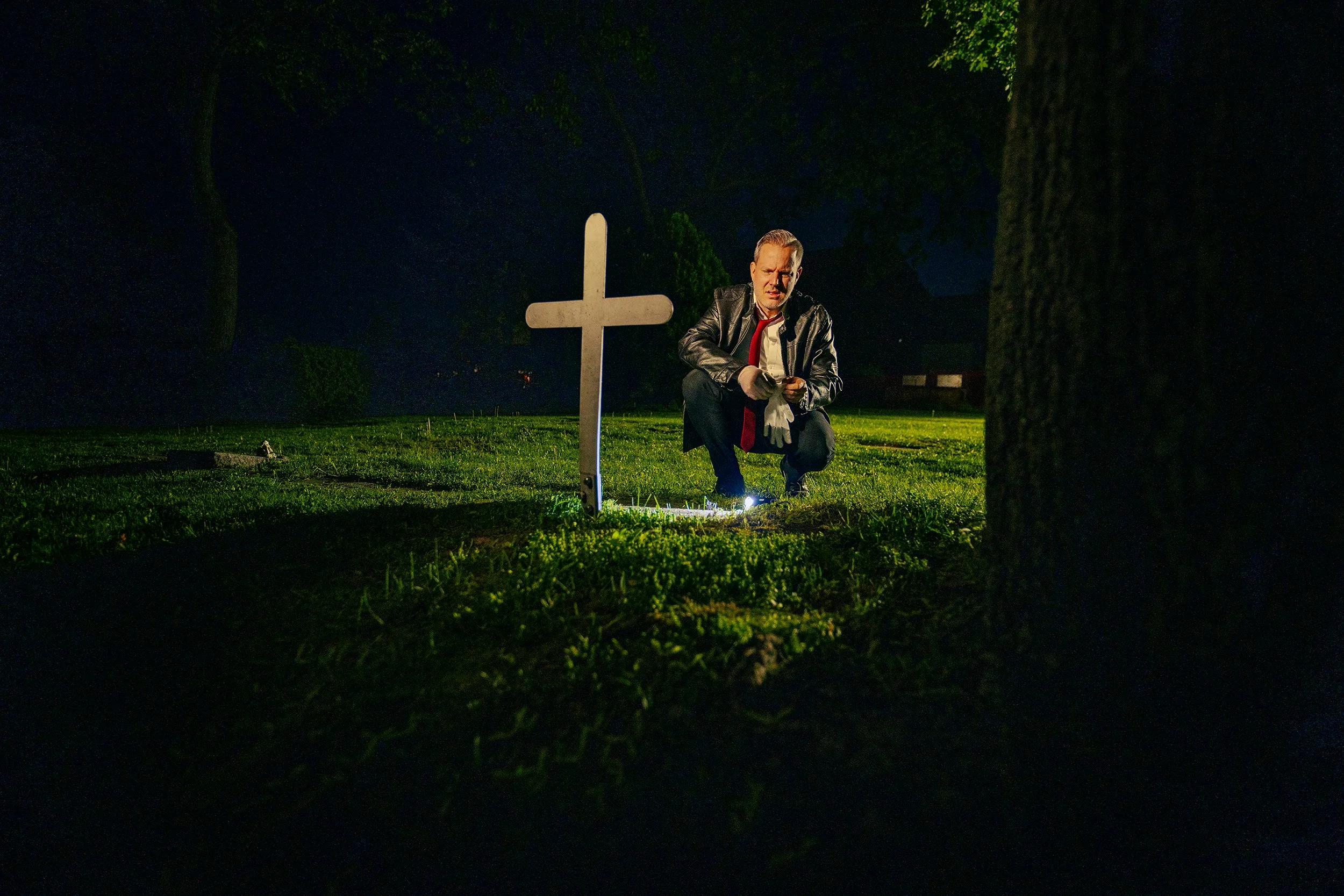 A man in a leather jacket and white gloves kneels on the grass at night near a white cross grave marker, illuminated by a light on the ground, with trees in the background.