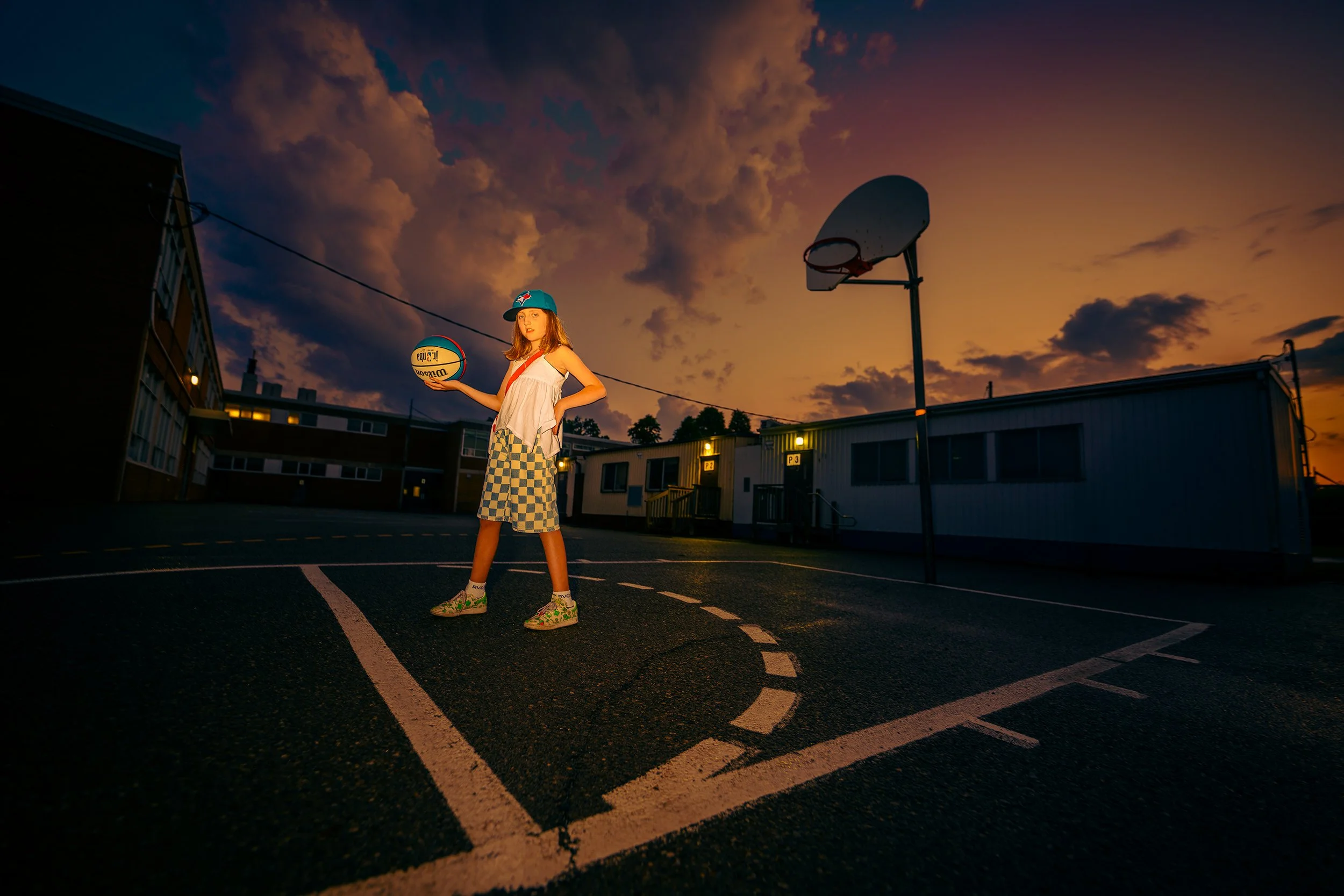 A young girl standing on an empty basketball court during sunset, holding a basketball and wearing a blue cap, striped shorts, and decorated shoes, with a sky filled with clouds in the background.