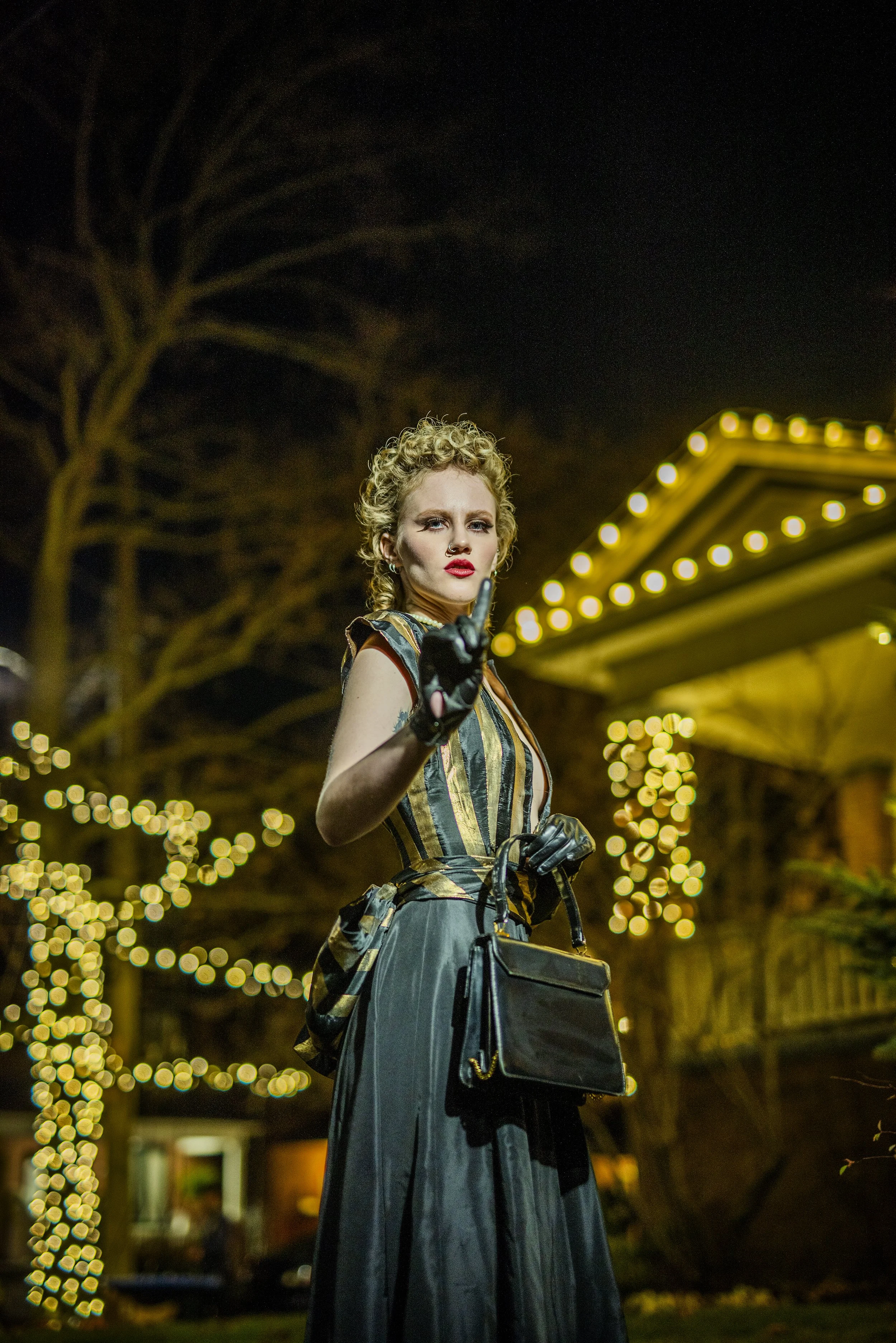 A woman standing outdoors at night near a lit structure and Christmas lights, wearing a striped dress, gloves, and holding a purse.