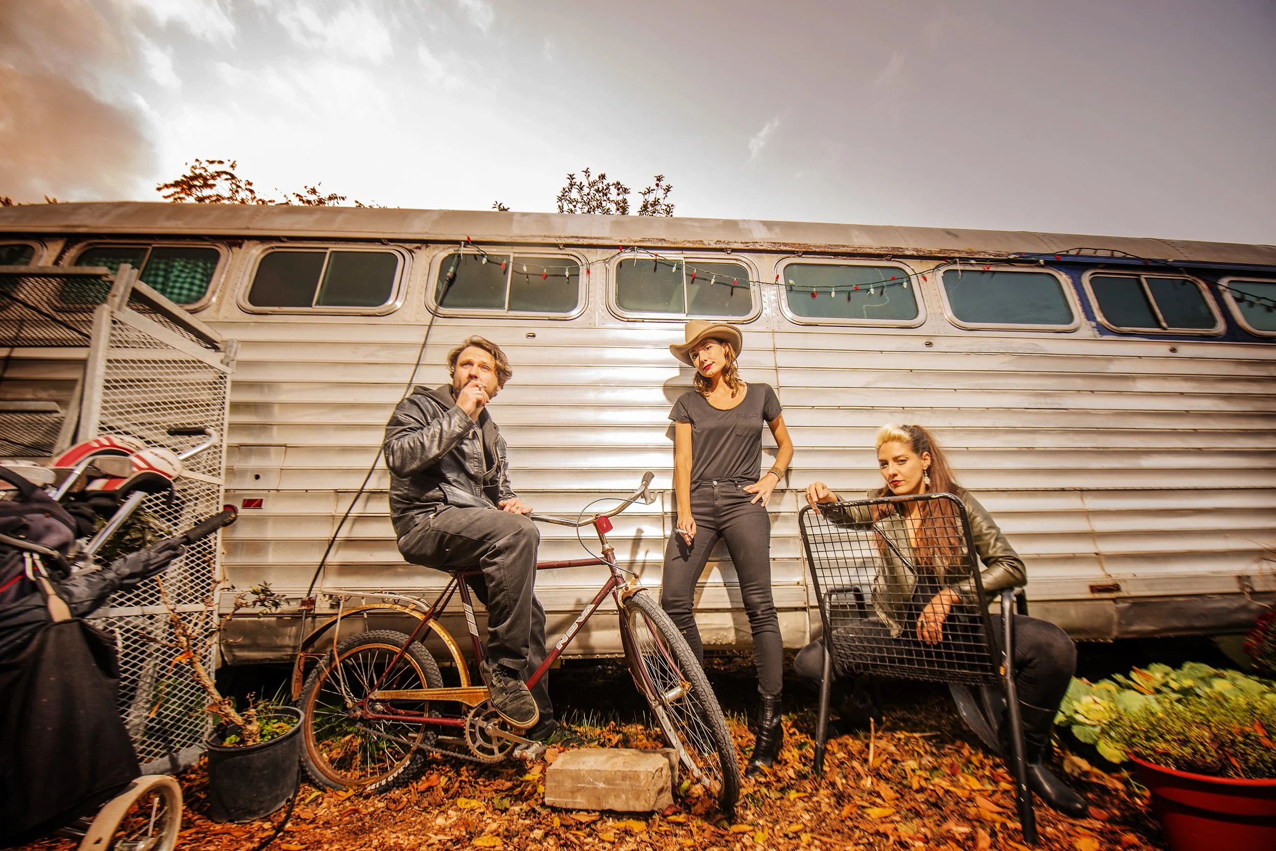 Three people are outdoors in front of a metallic trailer, with a cloudy sky above. One man is sitting on a bicycle, another woman is standing, and a third woman is crouching with a shopping cart. There are bikes and bags around them, and fallen autum