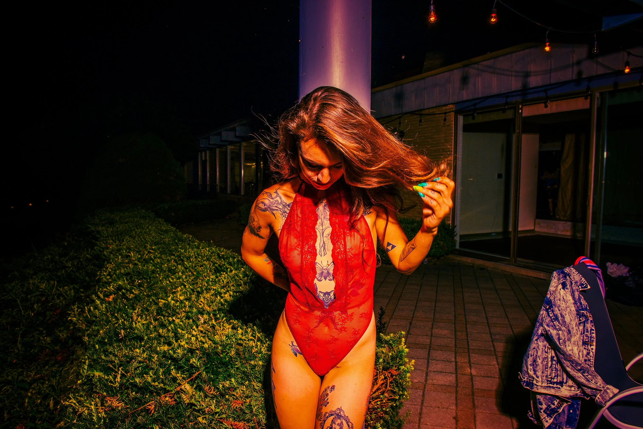 A woman with long hair wearing a red lace dress showing tattoos on her shoulders and legs, standing outdoors at night near a building with string lights, with her head bowed and hair flowing.