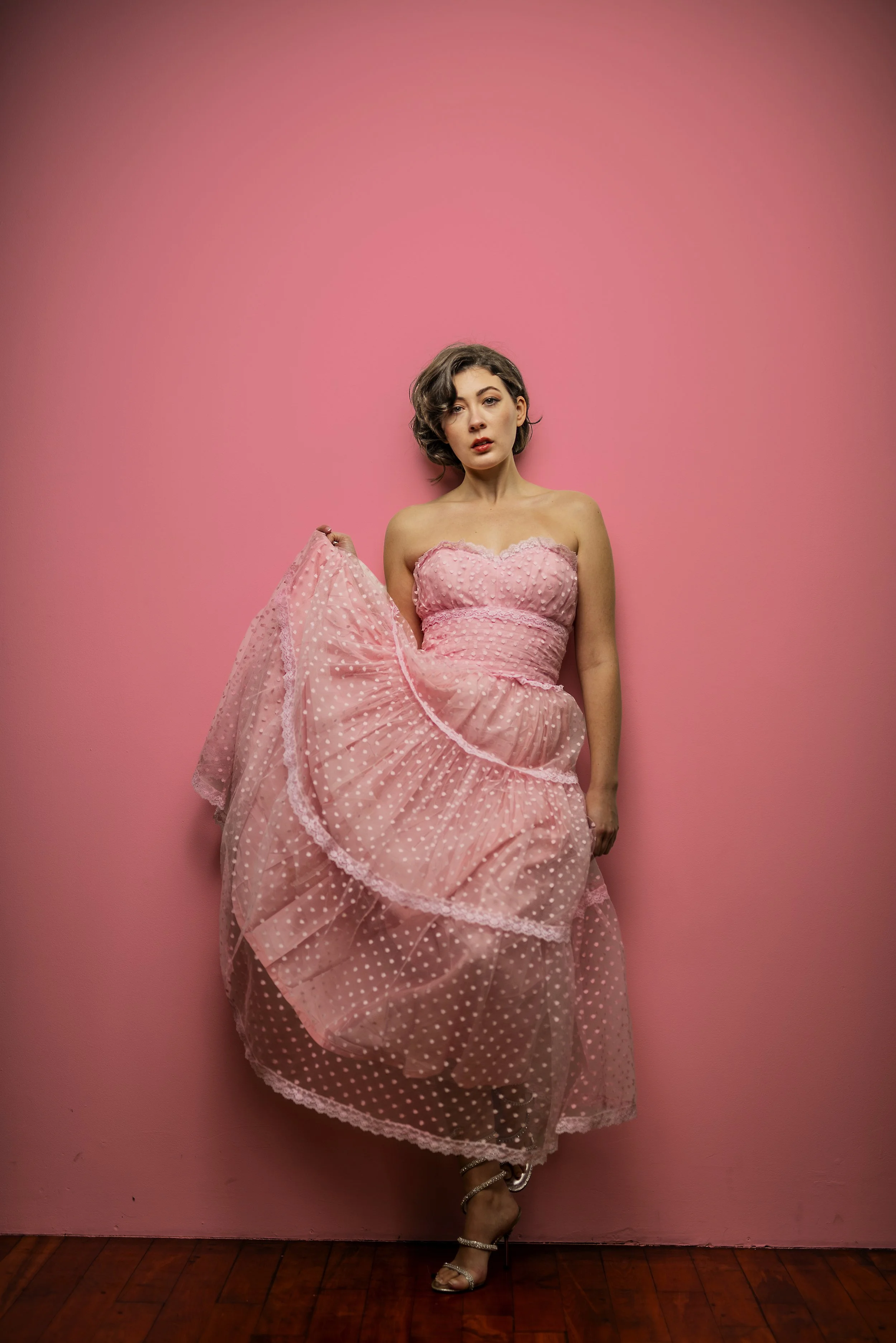 A woman wearing a strapless pink polka dot dress with ruffles, standing against a pink wall, holding up part of her dress, with one foot slightly raised, wearing silver high-heeled sandals with ankle straps.