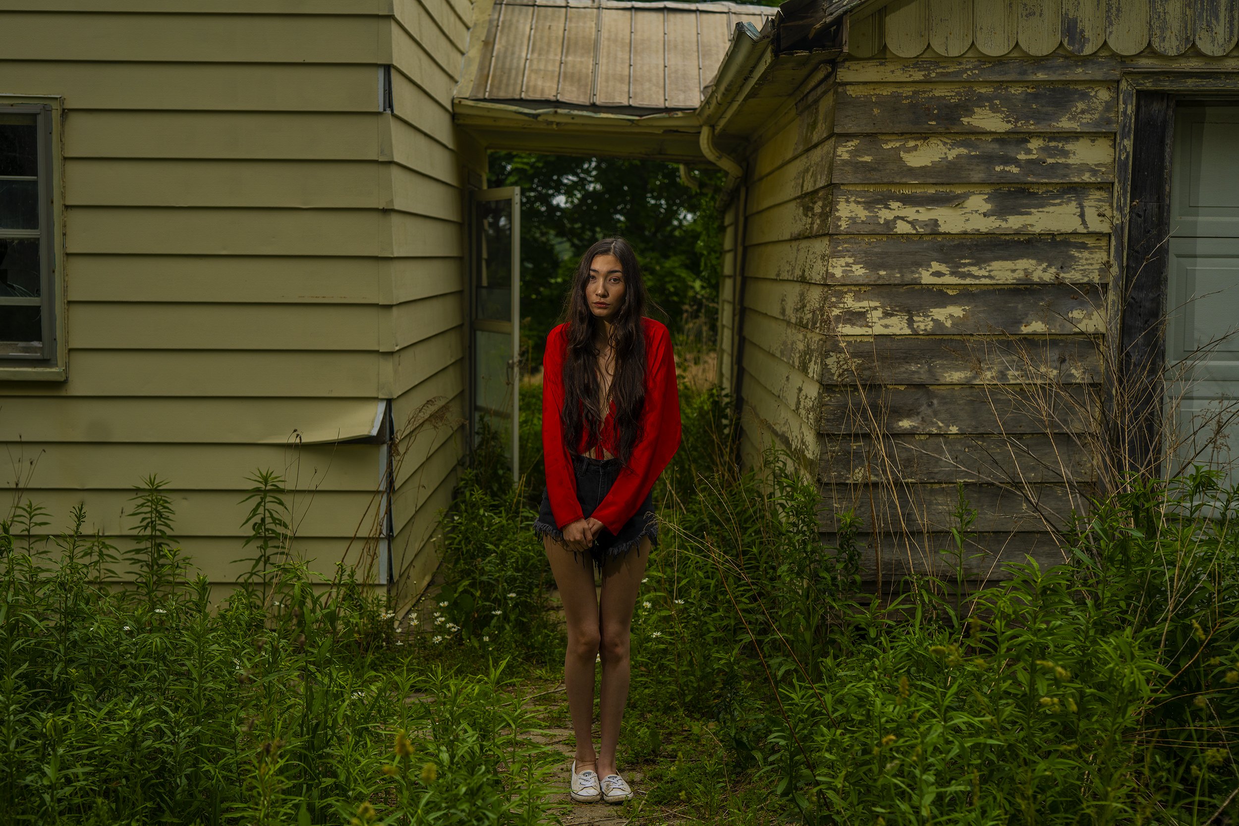 A young woman with long dark hair wearing a red jacket and black shorts standing outdoors on a pathway surrounded by tall green plants, with two old, weathered houses in the background.