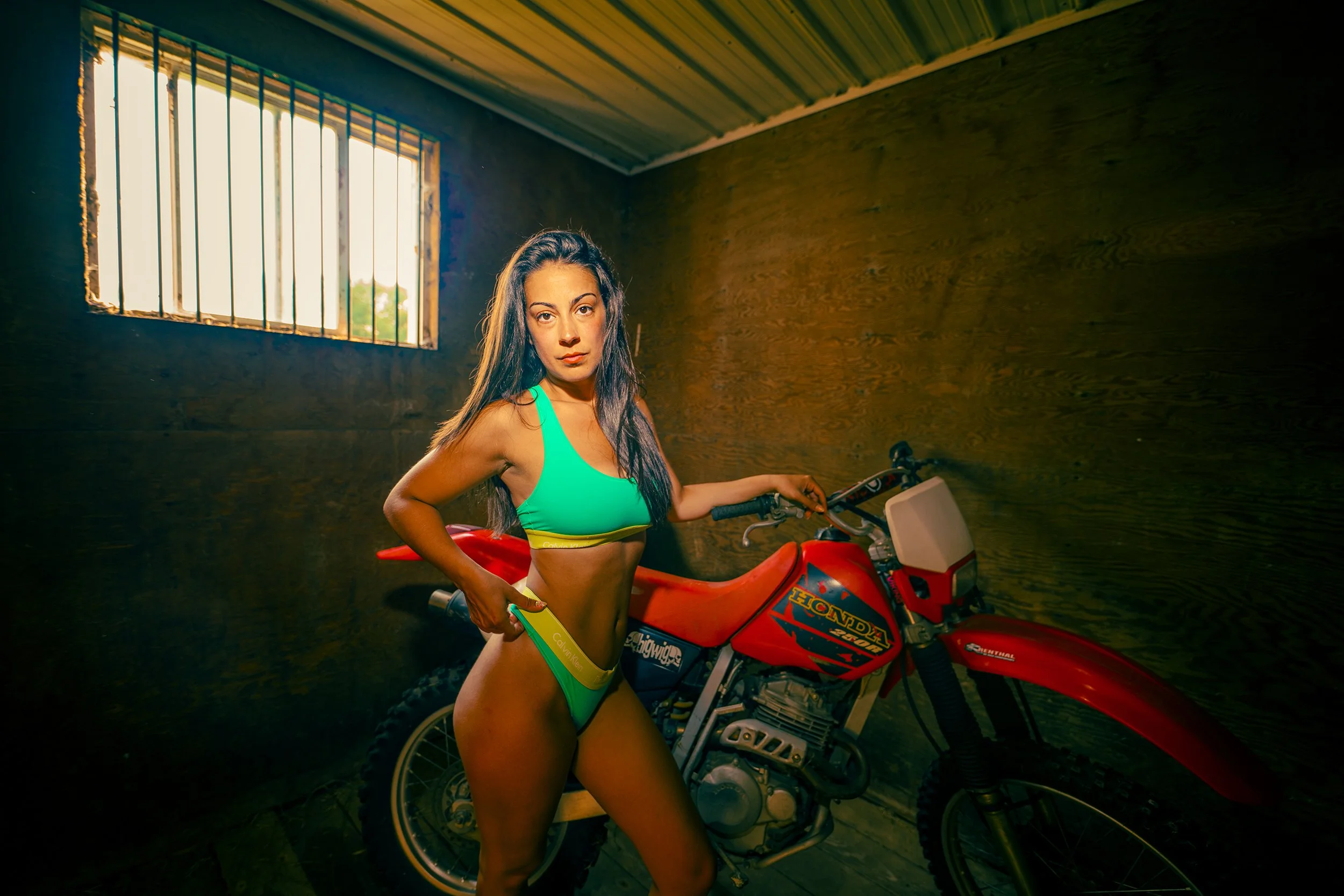Woman in green and yellow bikini holding a yellow paper near a red Honda motorcycle inside a dimly lit room with wooden walls and a barred window.