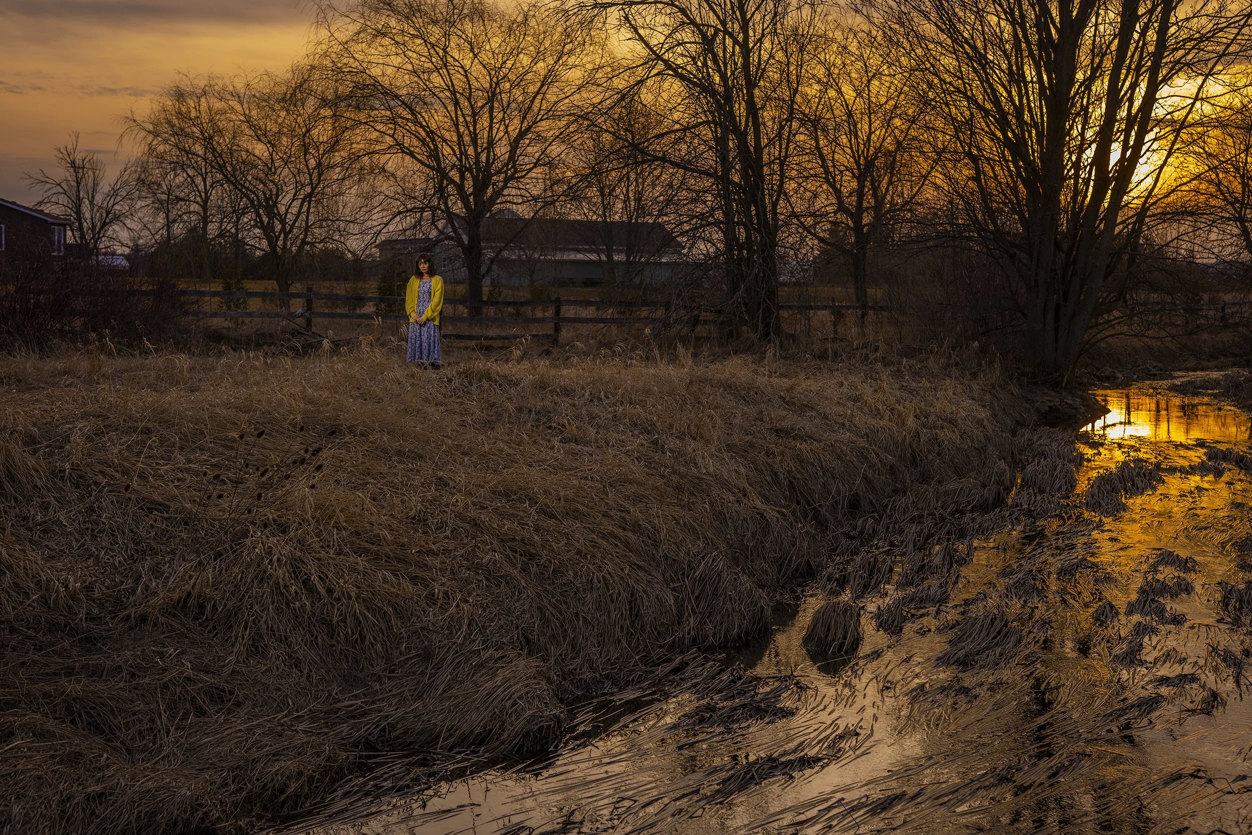 A woman in a yellow jacket standing on the bank of a creek during sunset with leafless trees and houses in the background.