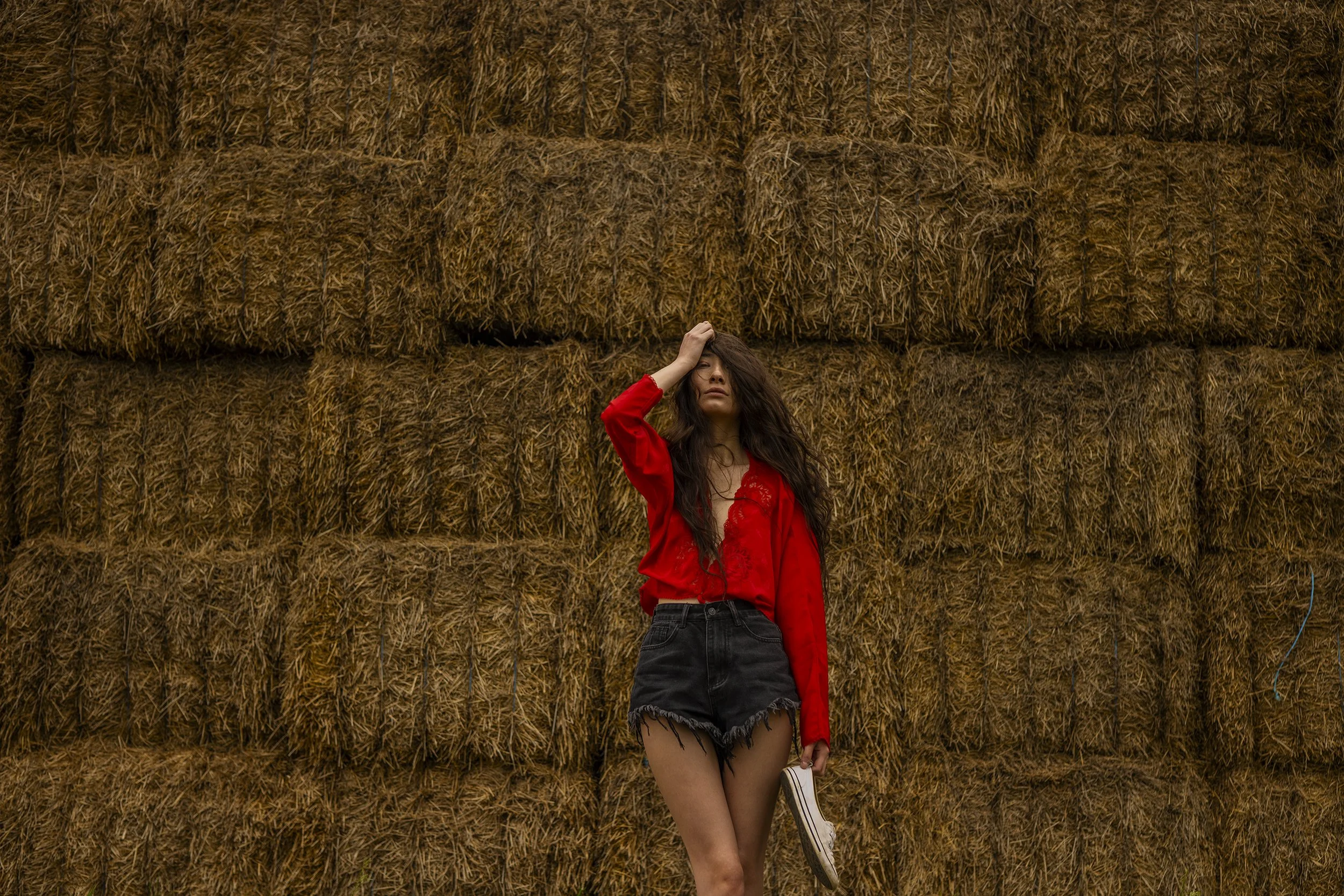 Young woman in red blouse and black shorts standing in front of stacked hay bales, holding a pair of shoes and touching her hair.