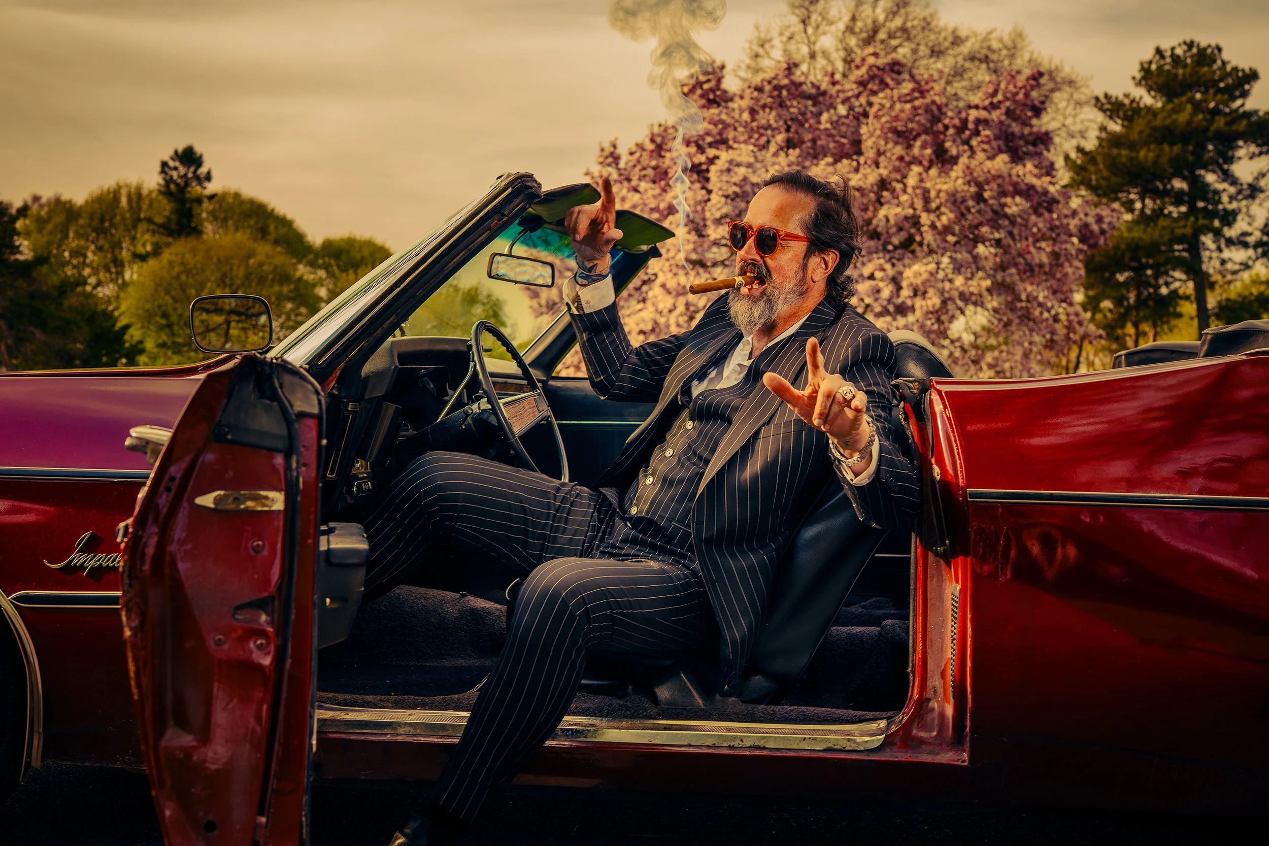 A man with sunglasses and a beard, dressed in a pinstripe suit, smoking a cigar, sitting in a vintage red convertible car during sunset with trees and a blossoming tree in the background.