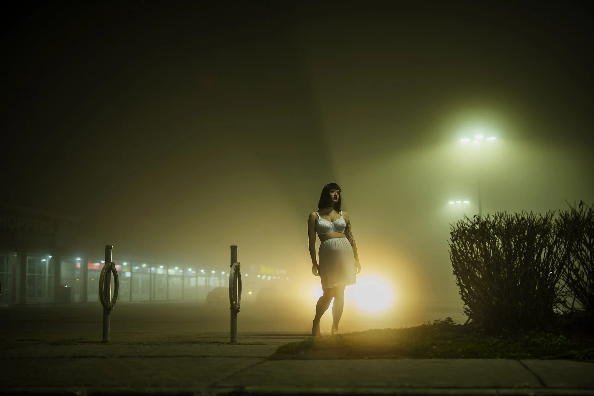 A woman standing alone on a foggy street at night, illuminated by a bright light source behind her, with streetlights and a bush visible in the background.